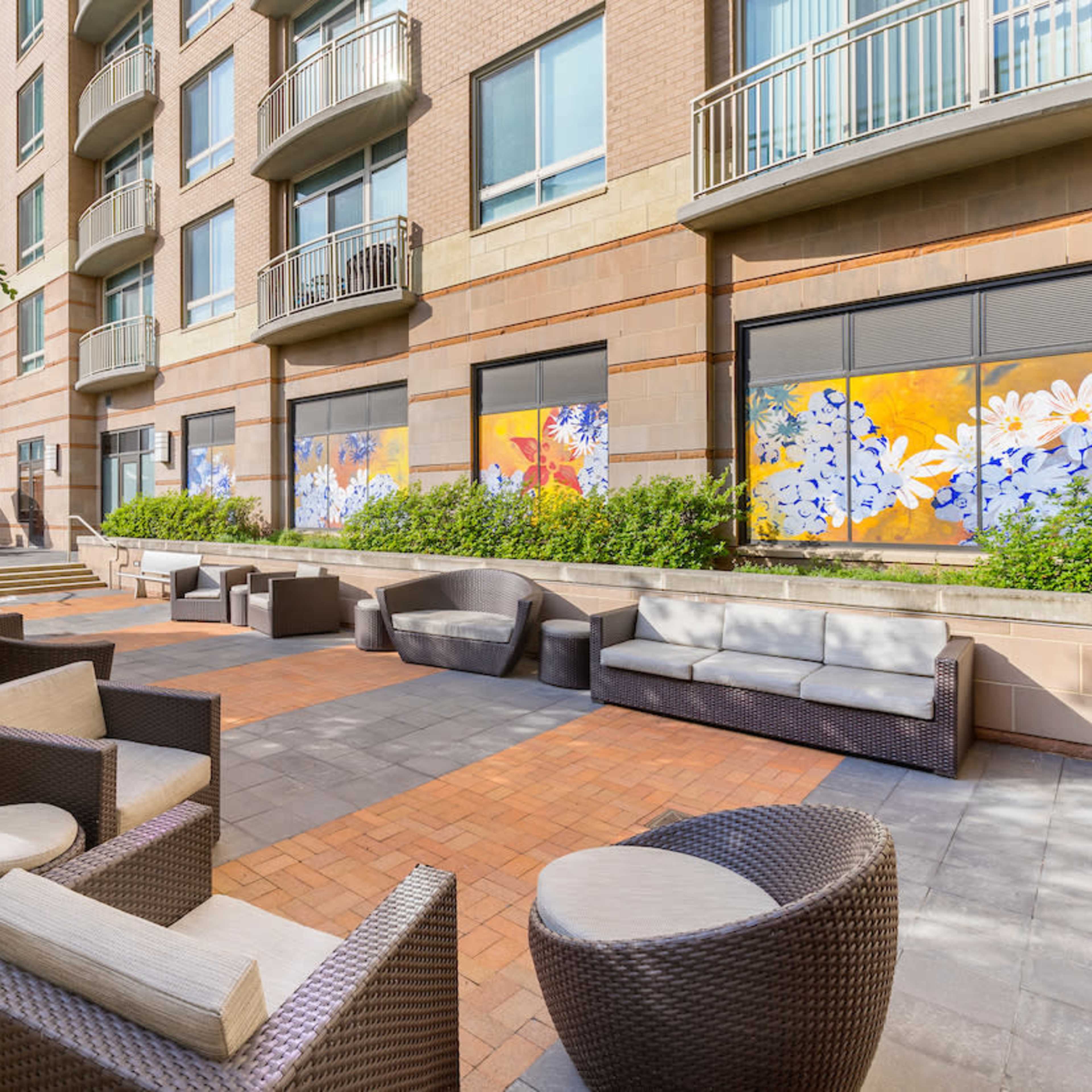 A patio area with modern furniture, including lounge chairs and tables, surrounded by greenery and a building with large windows.