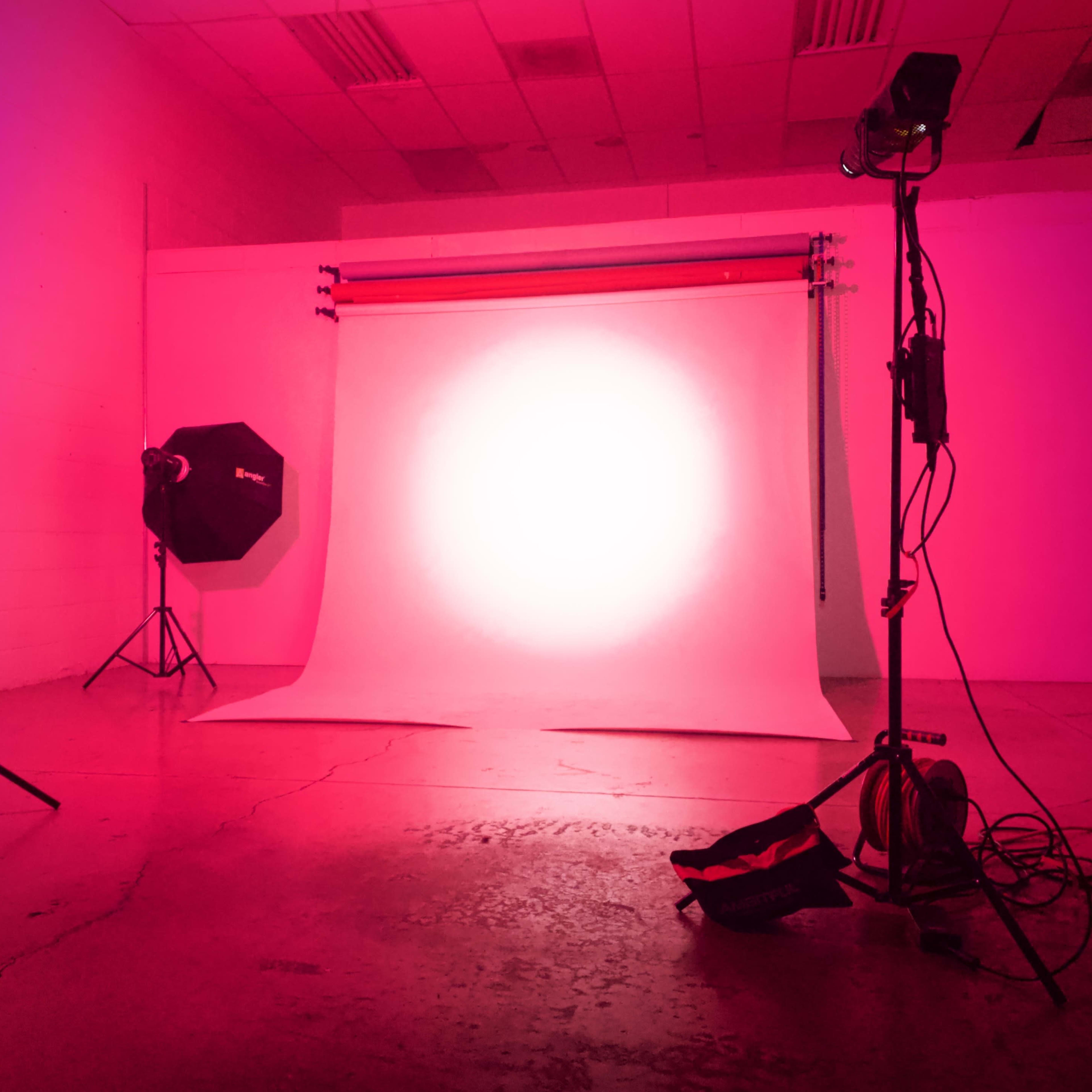 A photography studio with a pink backdrop, softbox, and lighting equipment positioned around a brightly lit area.
