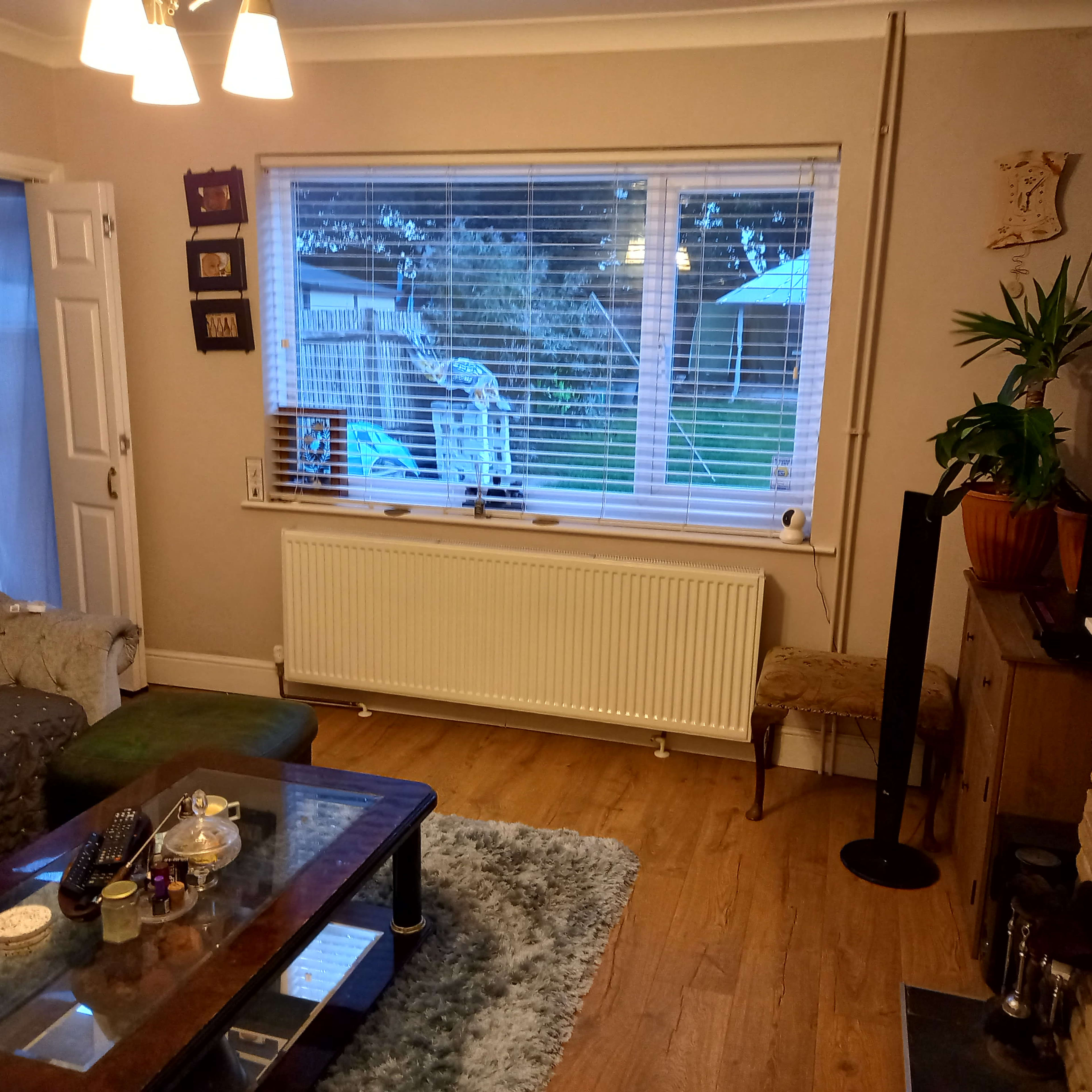 A cozy living room with a grey sofa, a coffee table displaying items, a large window with blinds, and a radiator underneath.