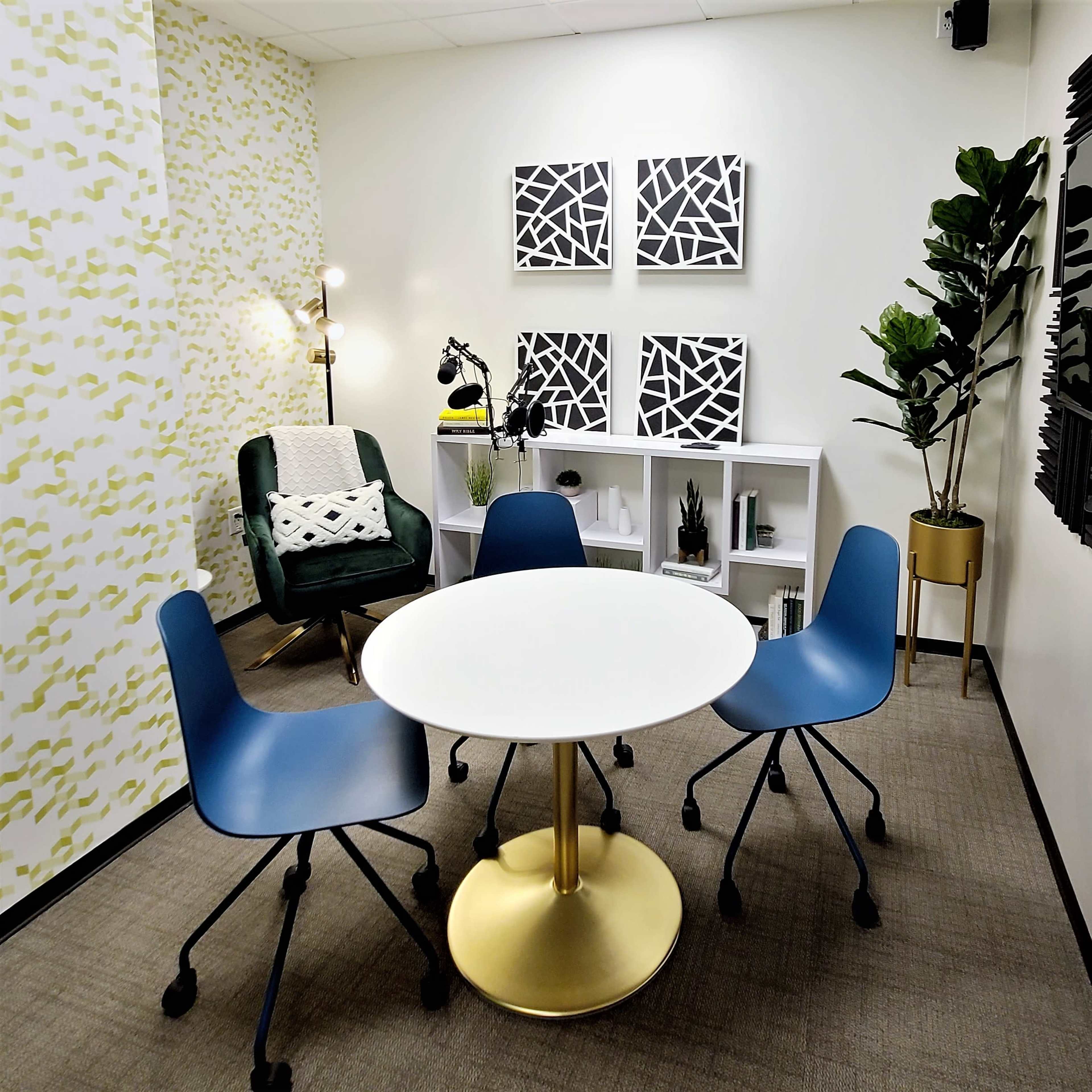 A small meeting room features a round white table surrounded by three blue chairs, with a green armchair, shelves, and black-and-white artwork on the walls.