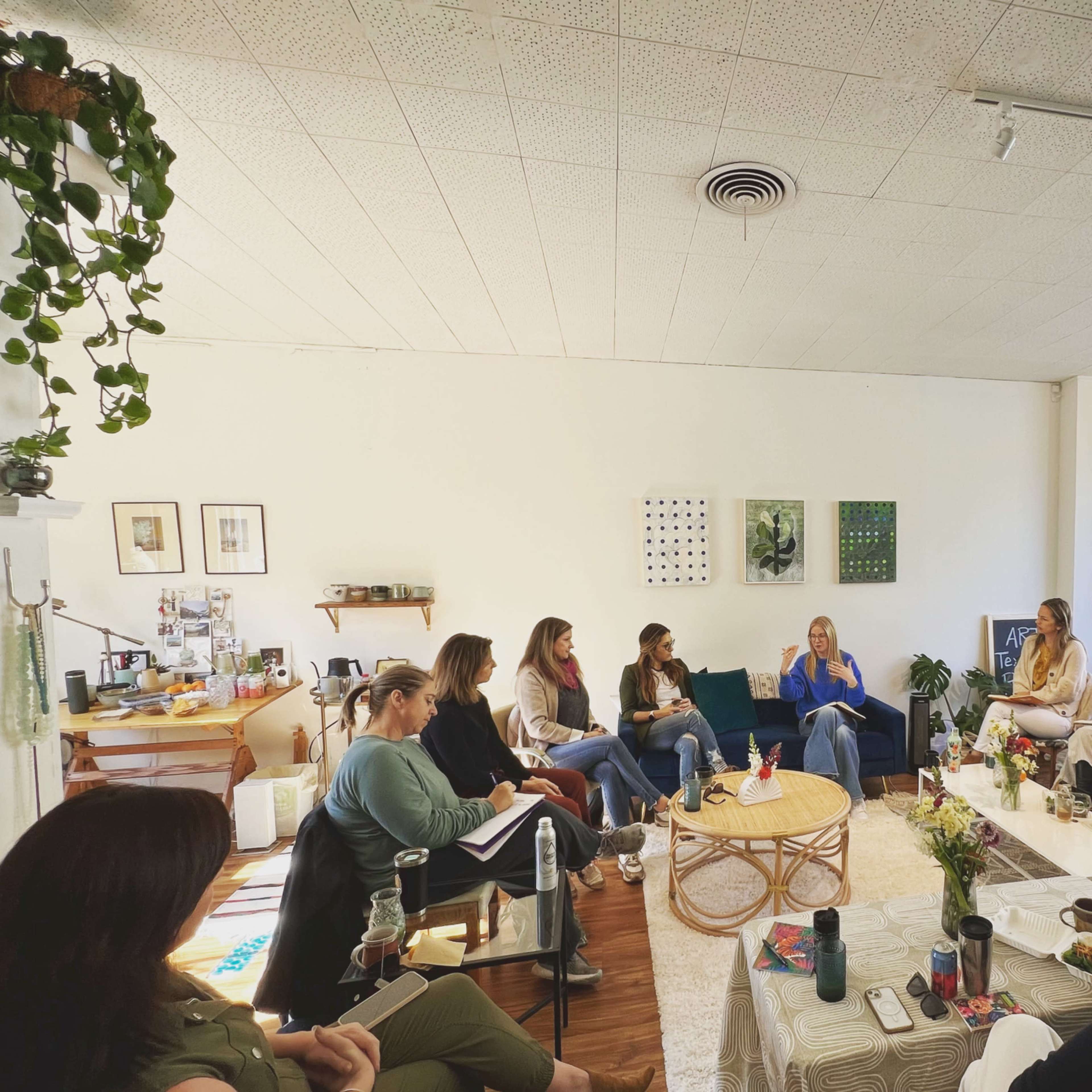 A group of women sits in a bright, open space, engaging in conversation while surrounded by plants and minimalist decor.