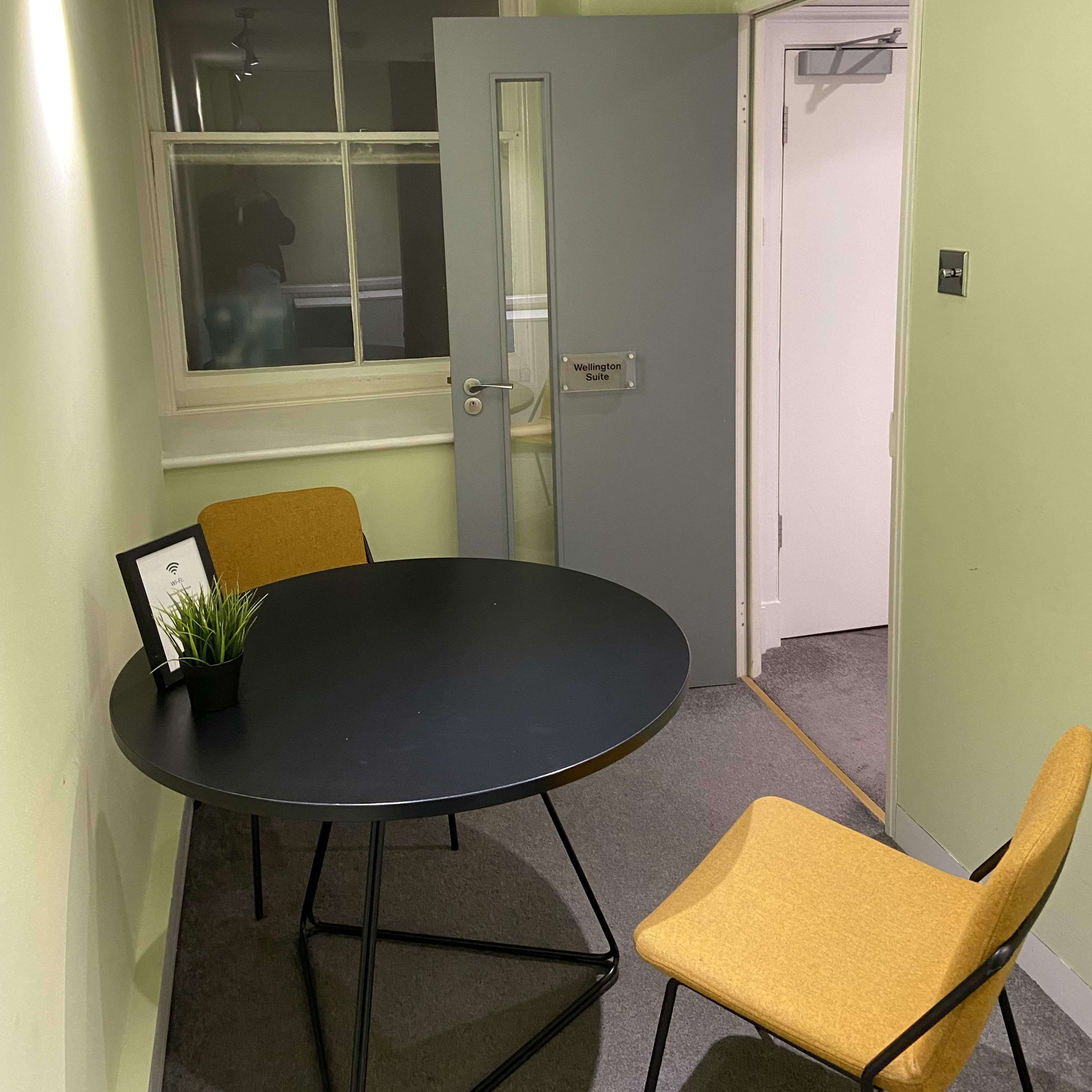 A small room with a round black table and two mustard-colored chairs, featuring a door partially open to an adjacent space.