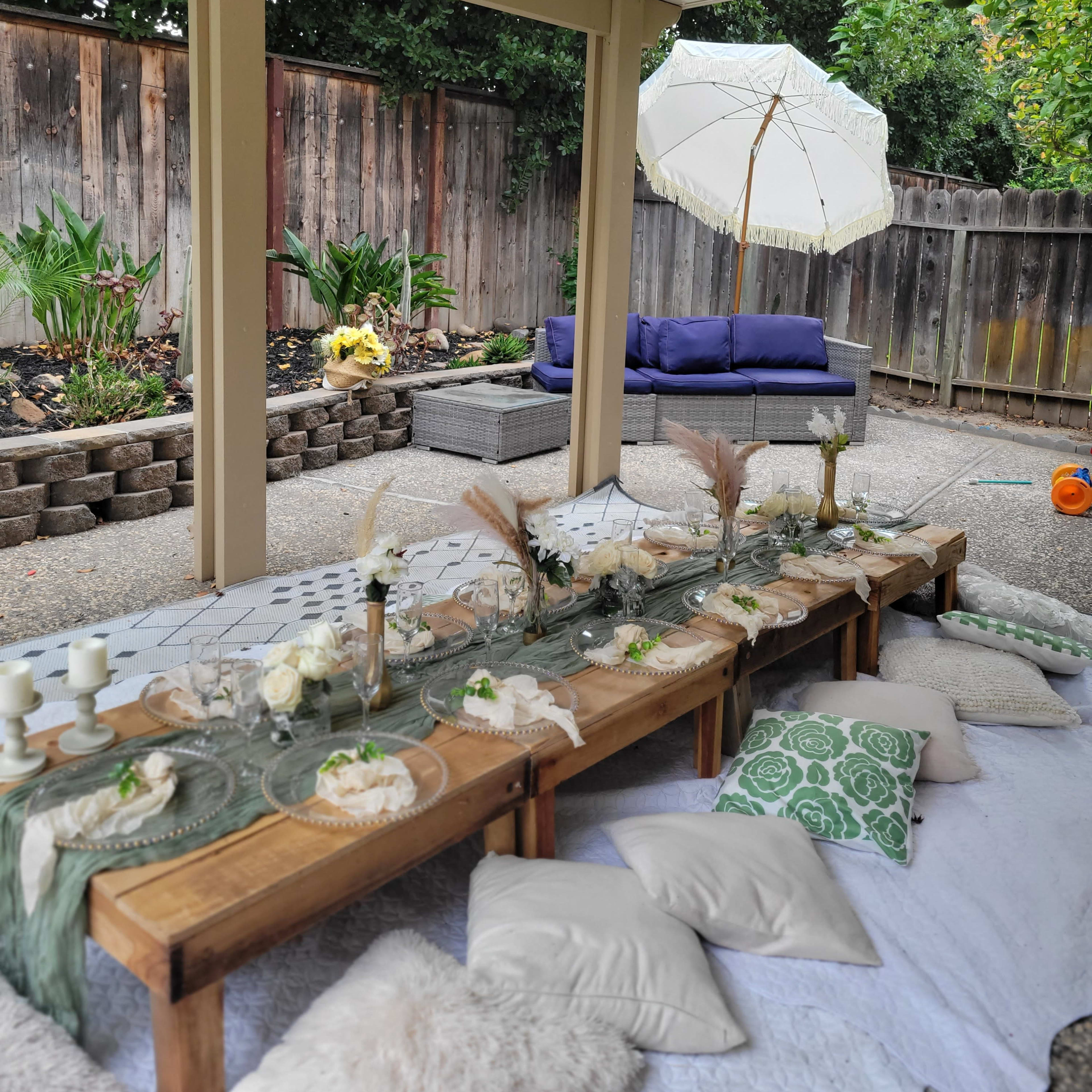A decorated outdoor seating area with a long wooden table set for a meal, surrounded by cushions and plants, and featuring an umbrella and a lounge area in the background.