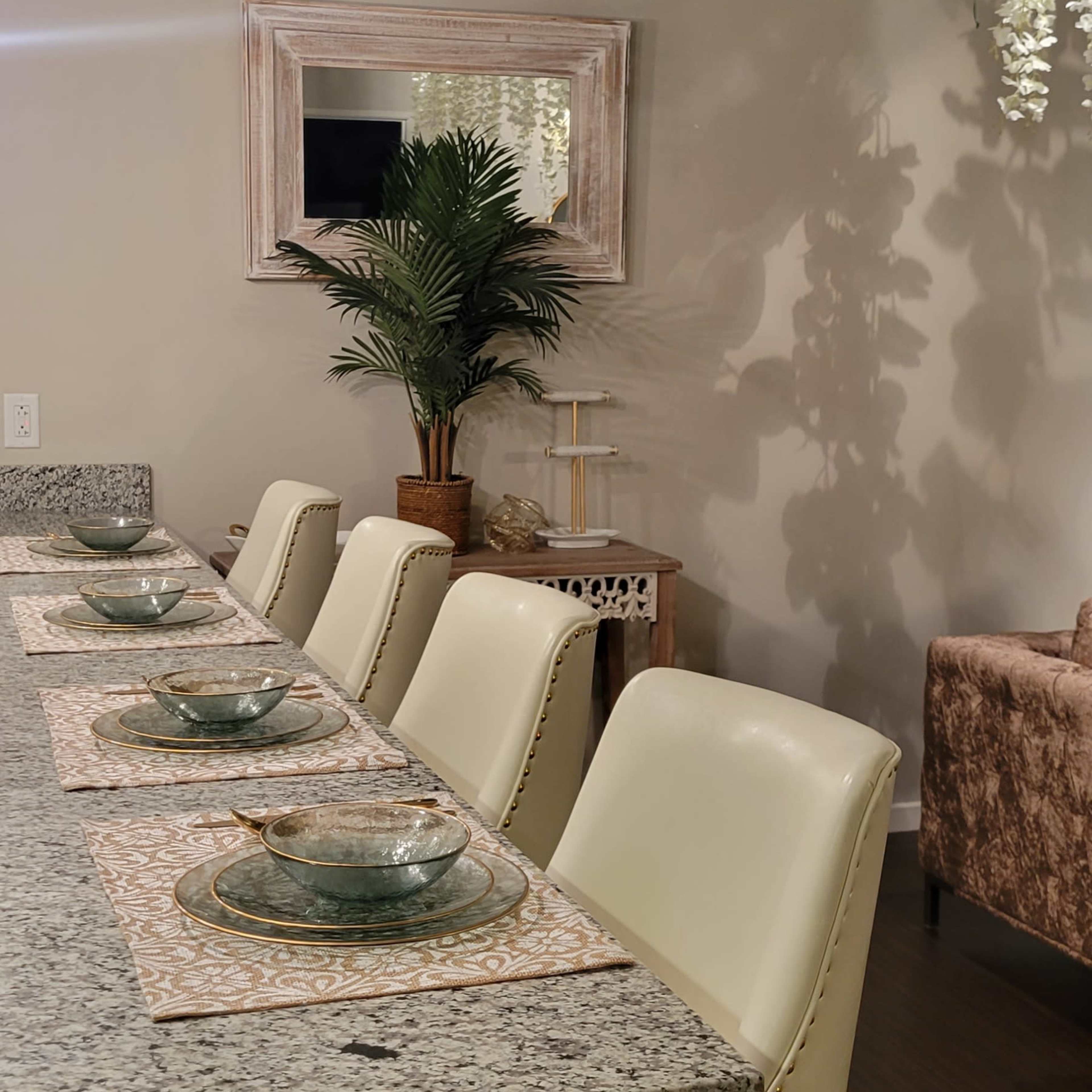 A modern dining area features a granite countertop with six white upholstered stools, elegant table settings, and a decorative plant in the background.