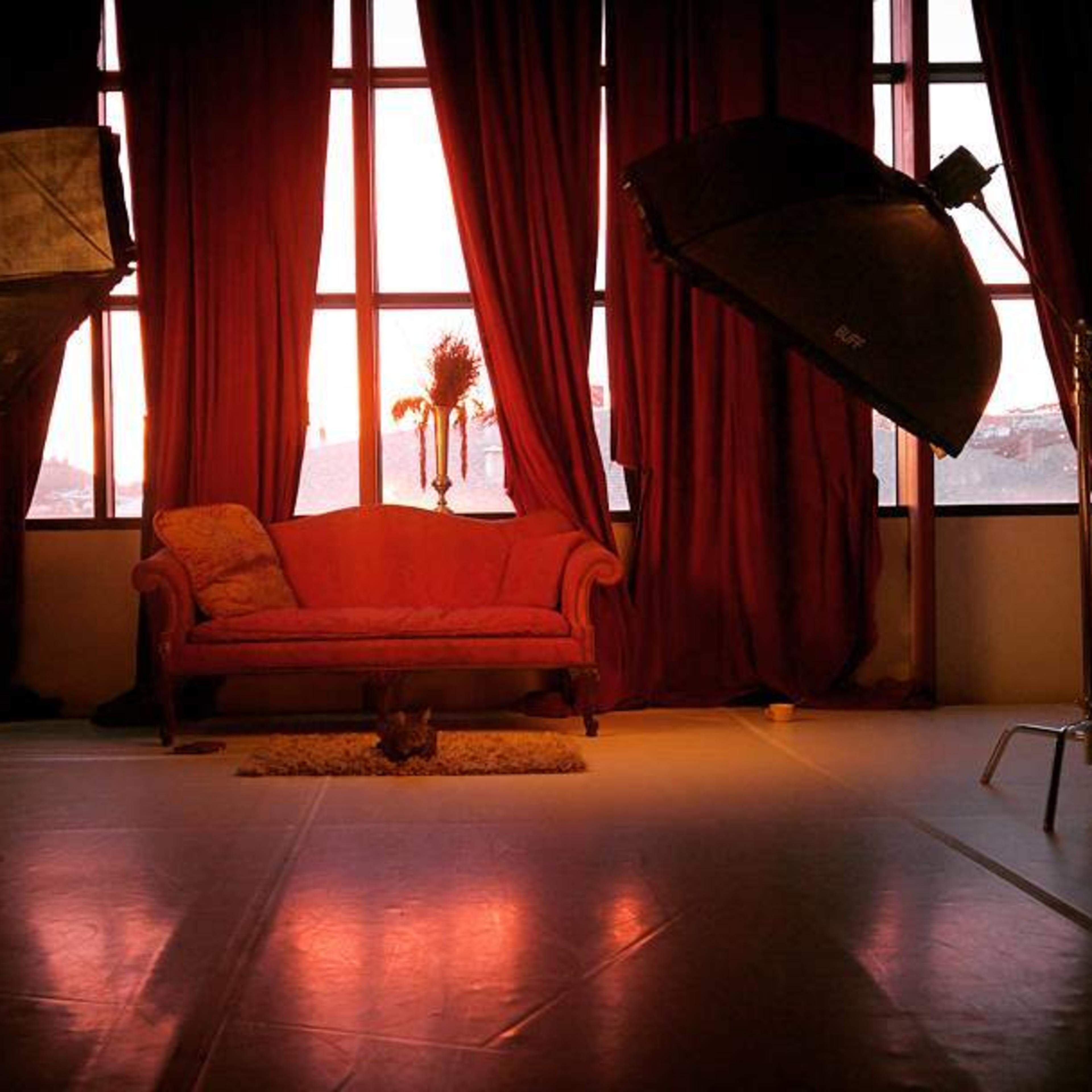 A pink couch is positioned in front of large windows, framed by red curtains, with two studio lights on either side.