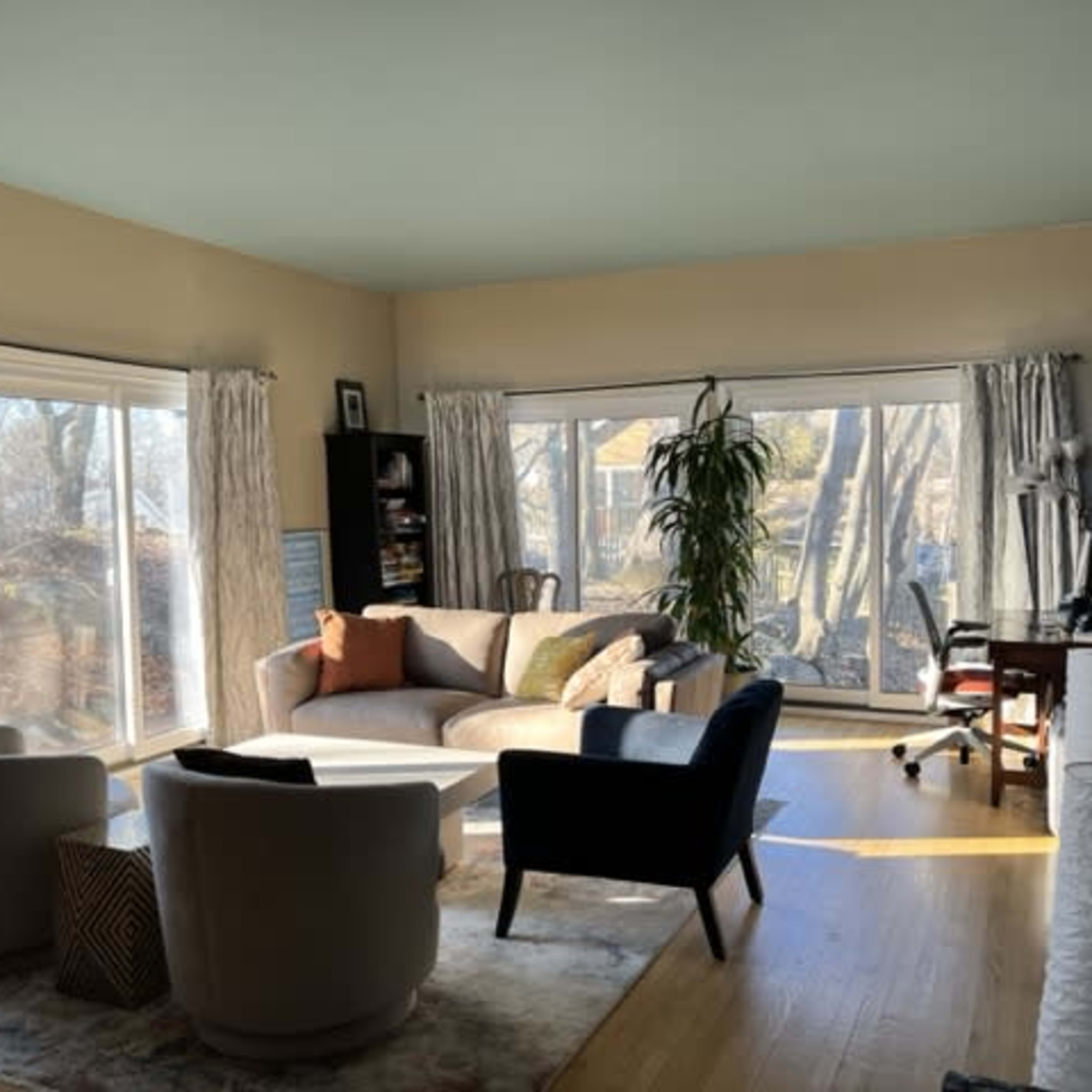 A bright living room features large windows, a curved sofa, two armchairs, and a desk near the corner.