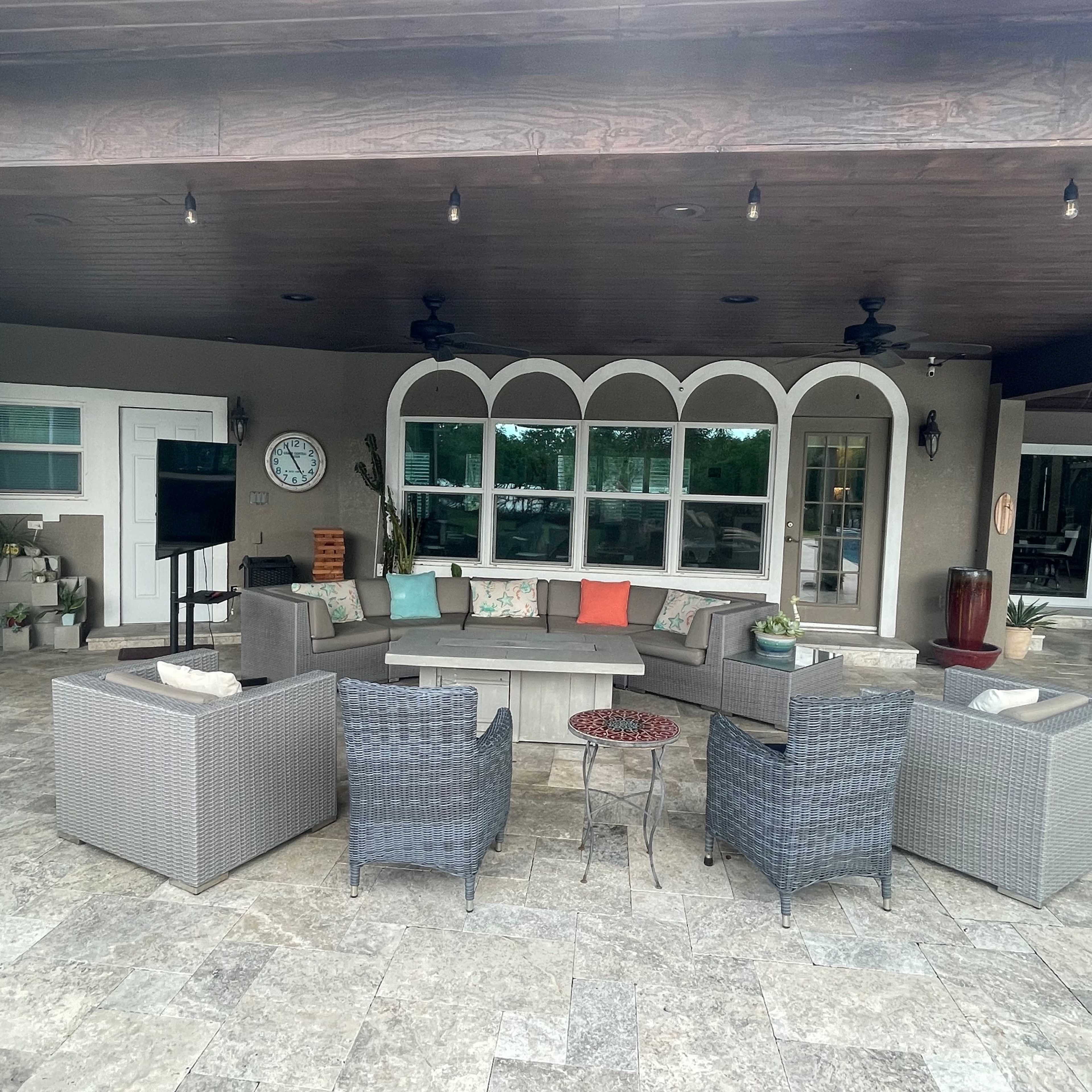 A spacious outdoor seating area with sectional couches and armchairs arranged around a small table, under a covered patio with decorative lighting.