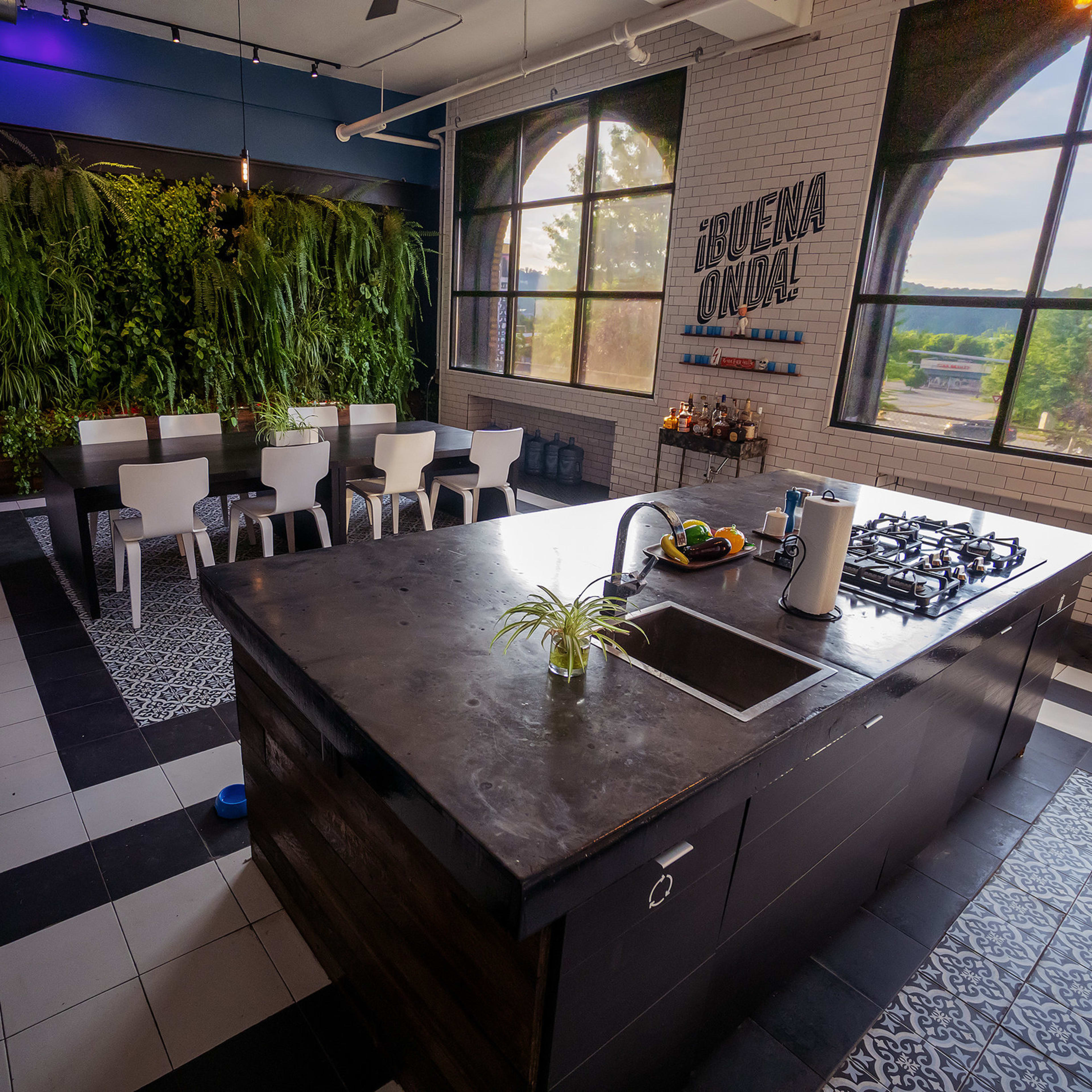 A modern kitchen features a large island with a sink and stove, surrounded by white chairs and a wall of greenery, alongside large windows that provide natural light.