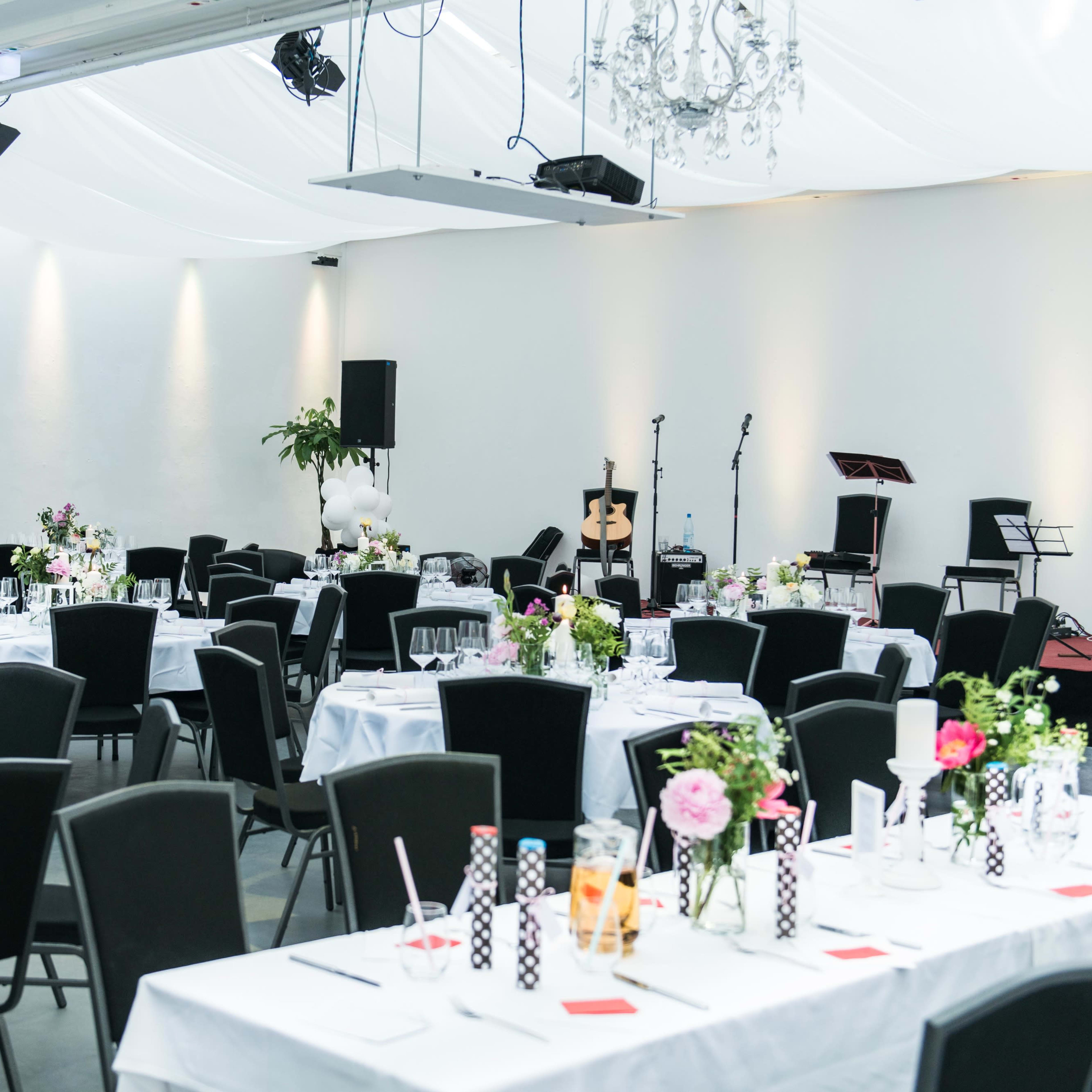 A banquet hall is set with elegantly arranged tables, each adorned with floral centerpieces and surrounded by black chairs, under soft lighting and decorative chandeliers.