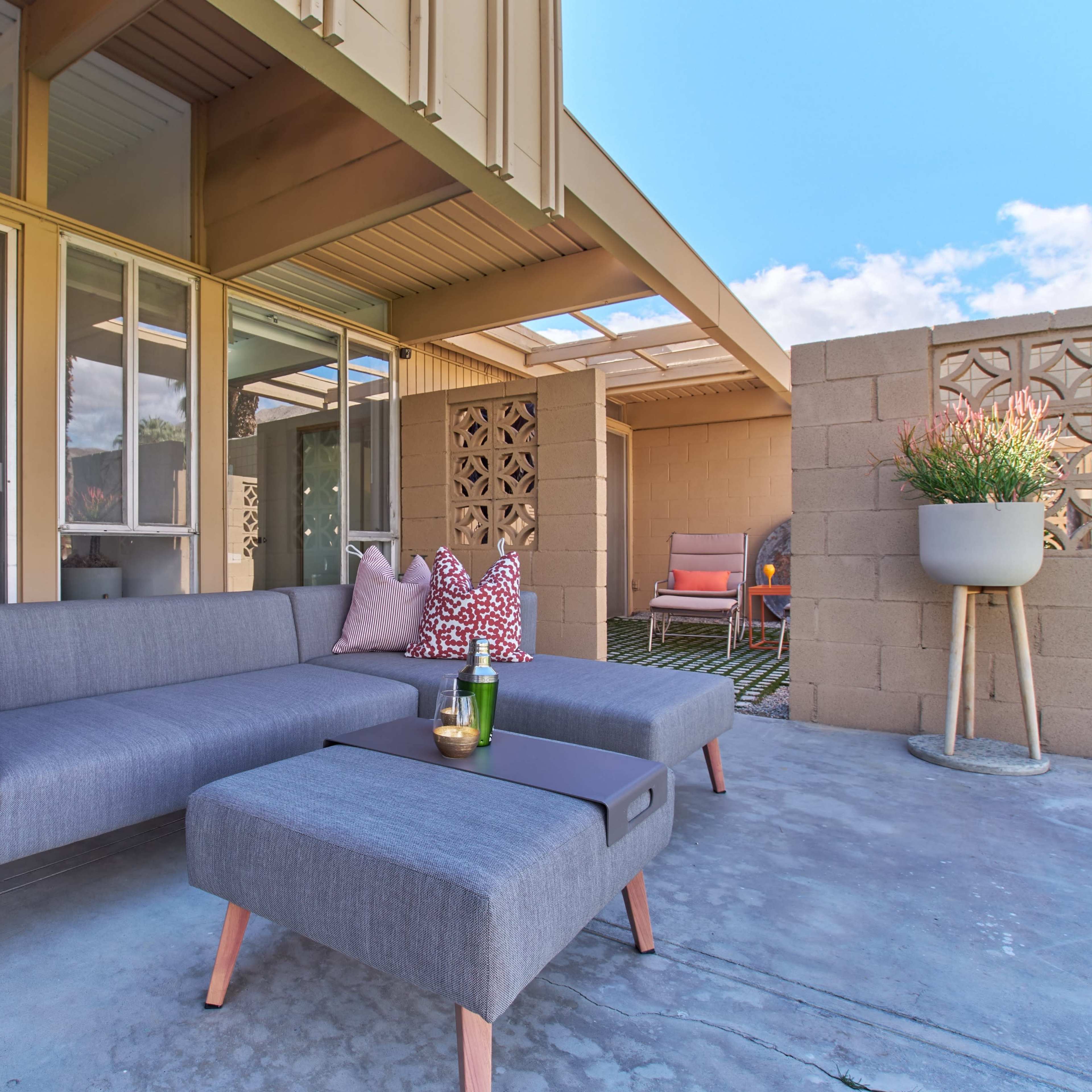 A gray sectional sofa and matching ottoman are set on a concrete patio, surrounded by large windows and a decorative block wall.
