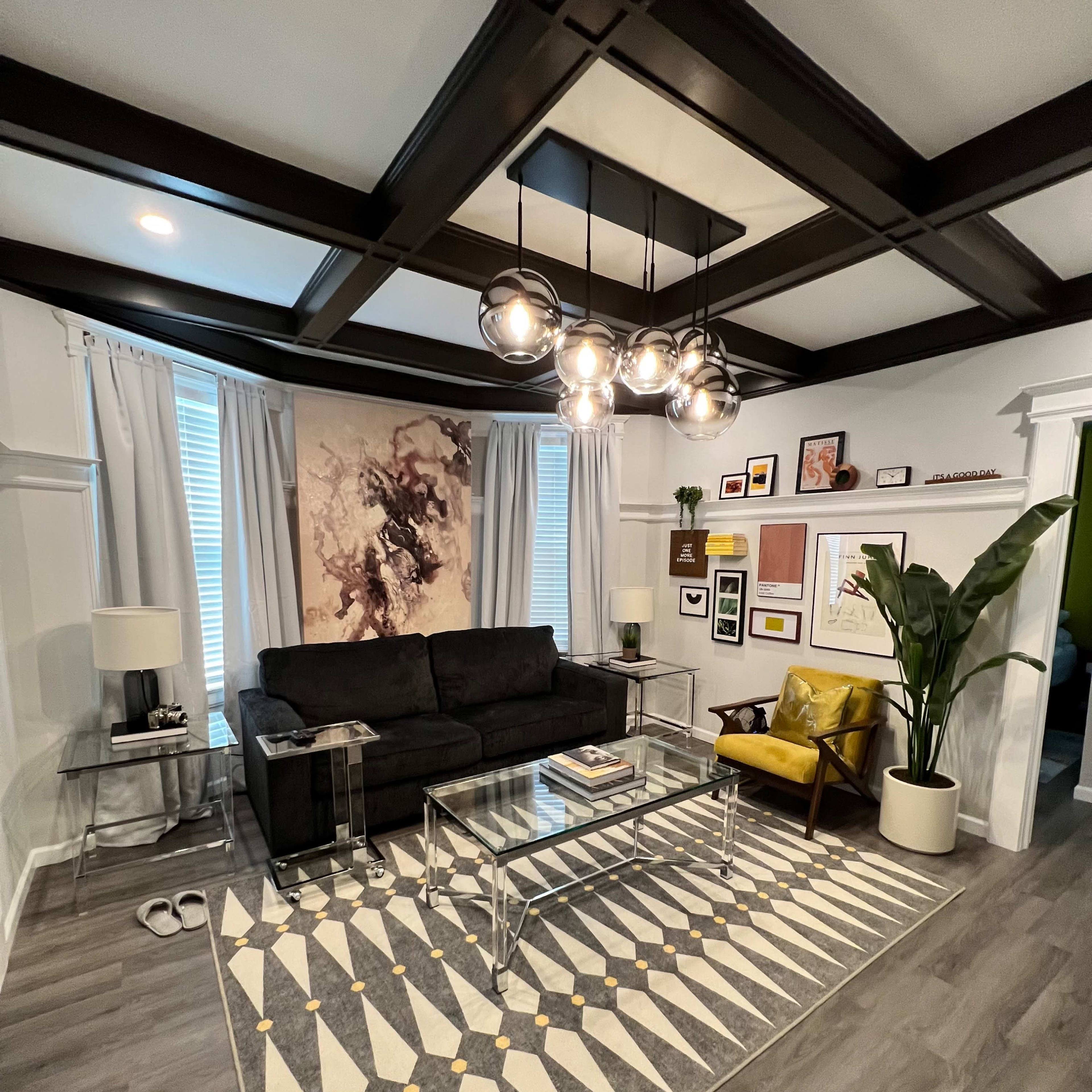 A modern living room featuring a black sofa, a yellow accent chair, a glass coffee table on a patterned rug, and various artworks framed on the walls.
