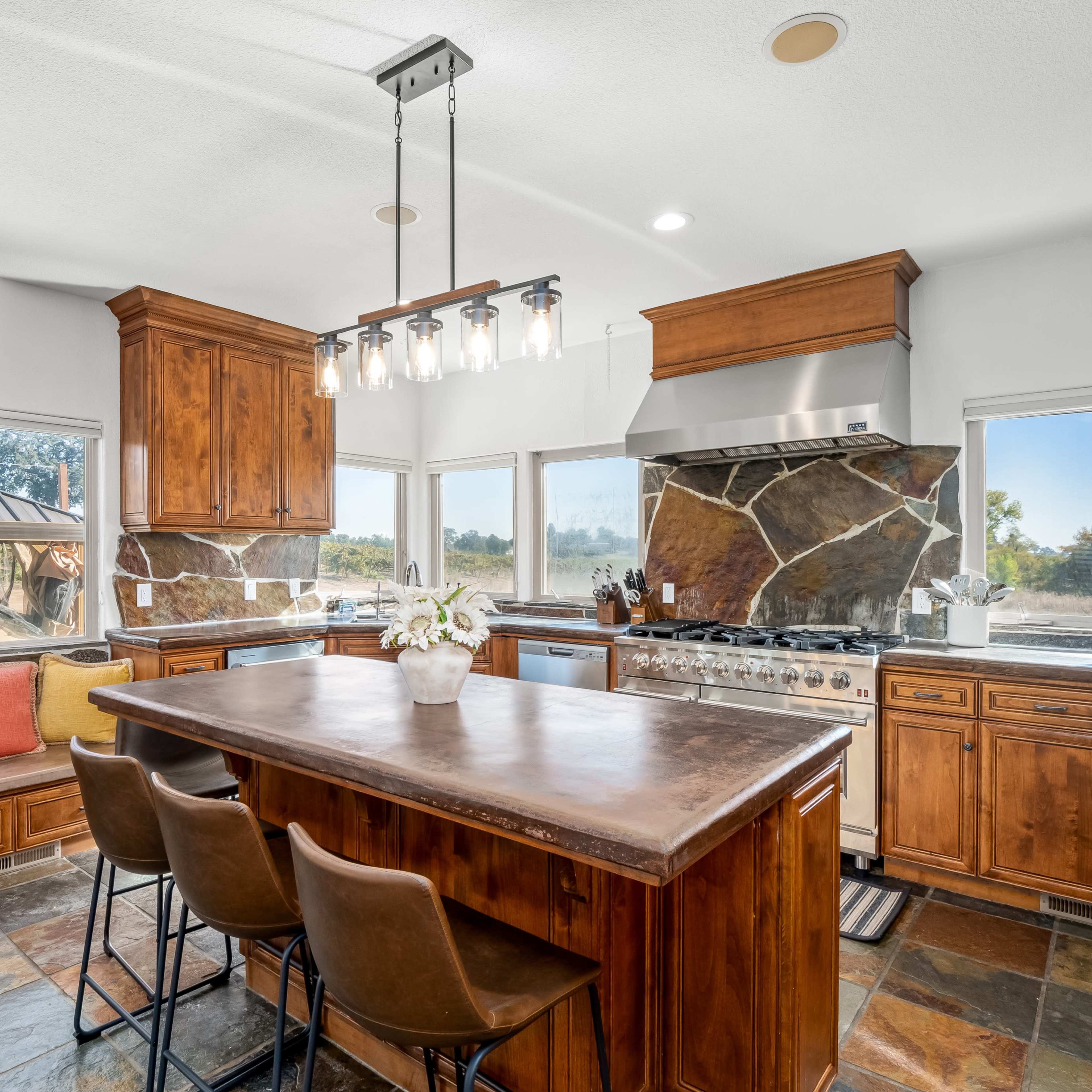 A modern kitchen features wooden cabinets, a large island with seating, and large windows offering a view of the outdoors.