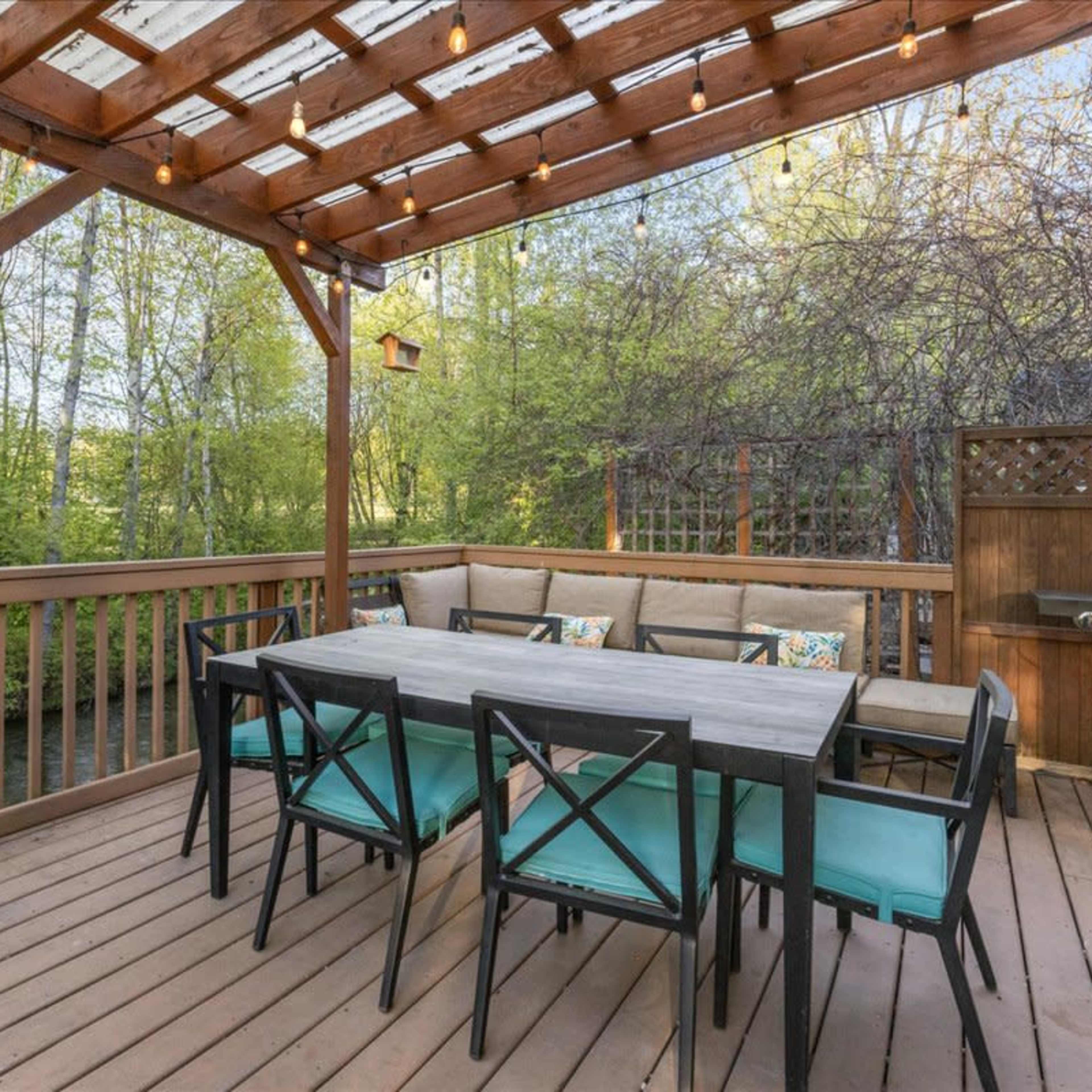 A wooden deck features a large dining table surrounded by chairs, with a grill and greenery visible in the background.