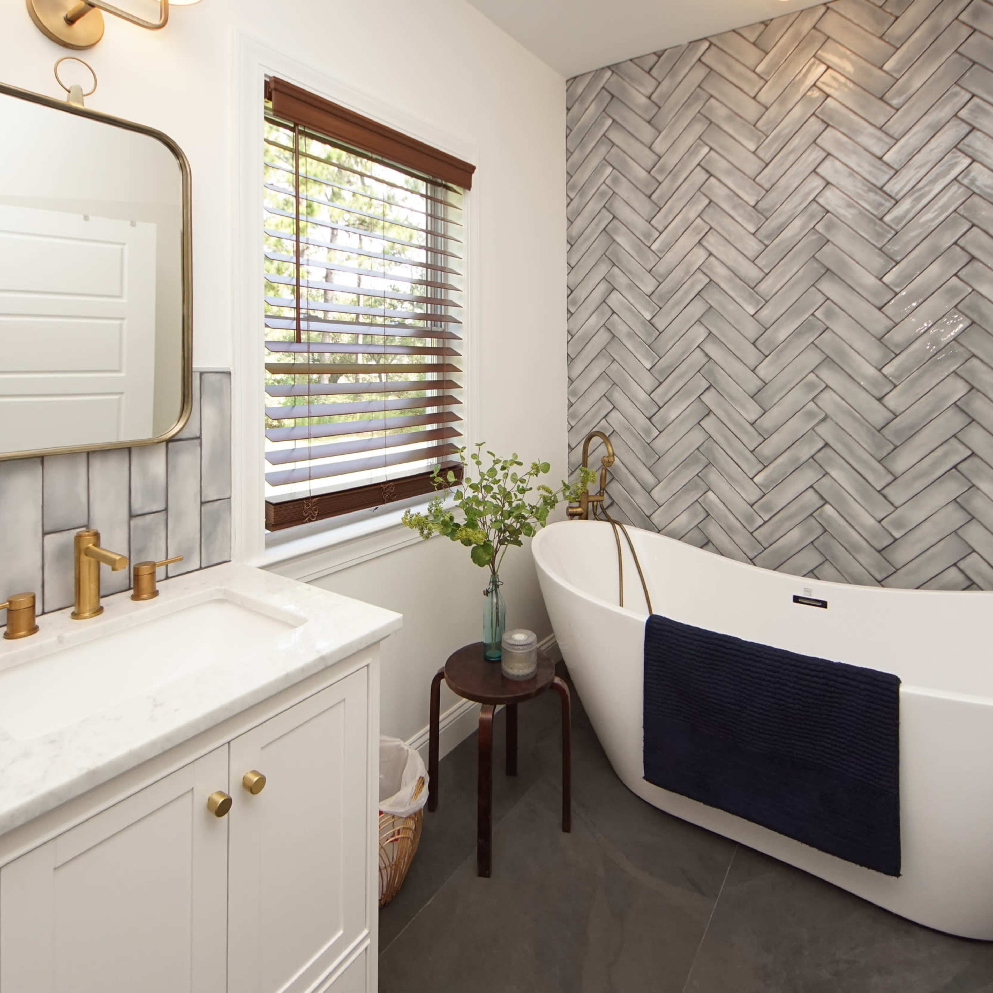 A bathroom featuring a freestanding tub, a herringbone-patterned tile wall, a sink with a mirror, and a window with wooden blinds.