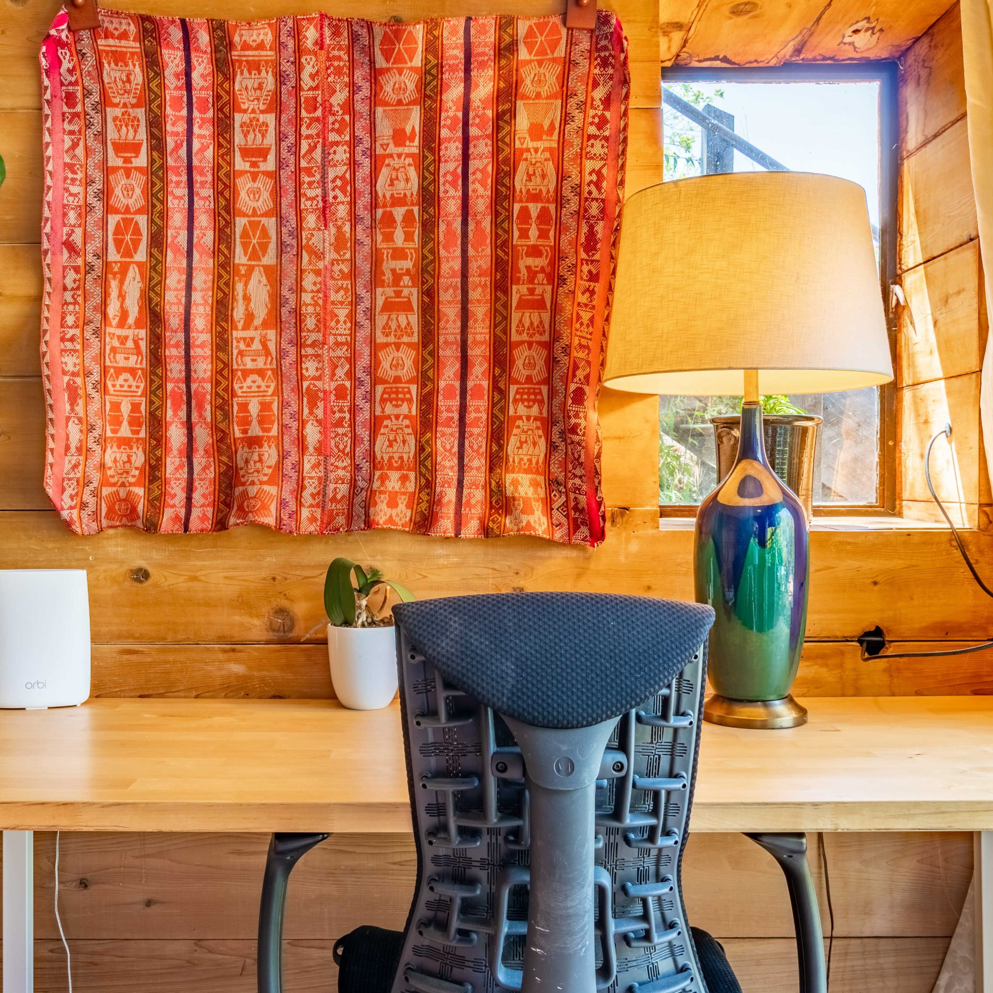 A wooden workspace featuring a desk with a chair, a decorative fabric hanging over a window, a lamp, and a small plant.