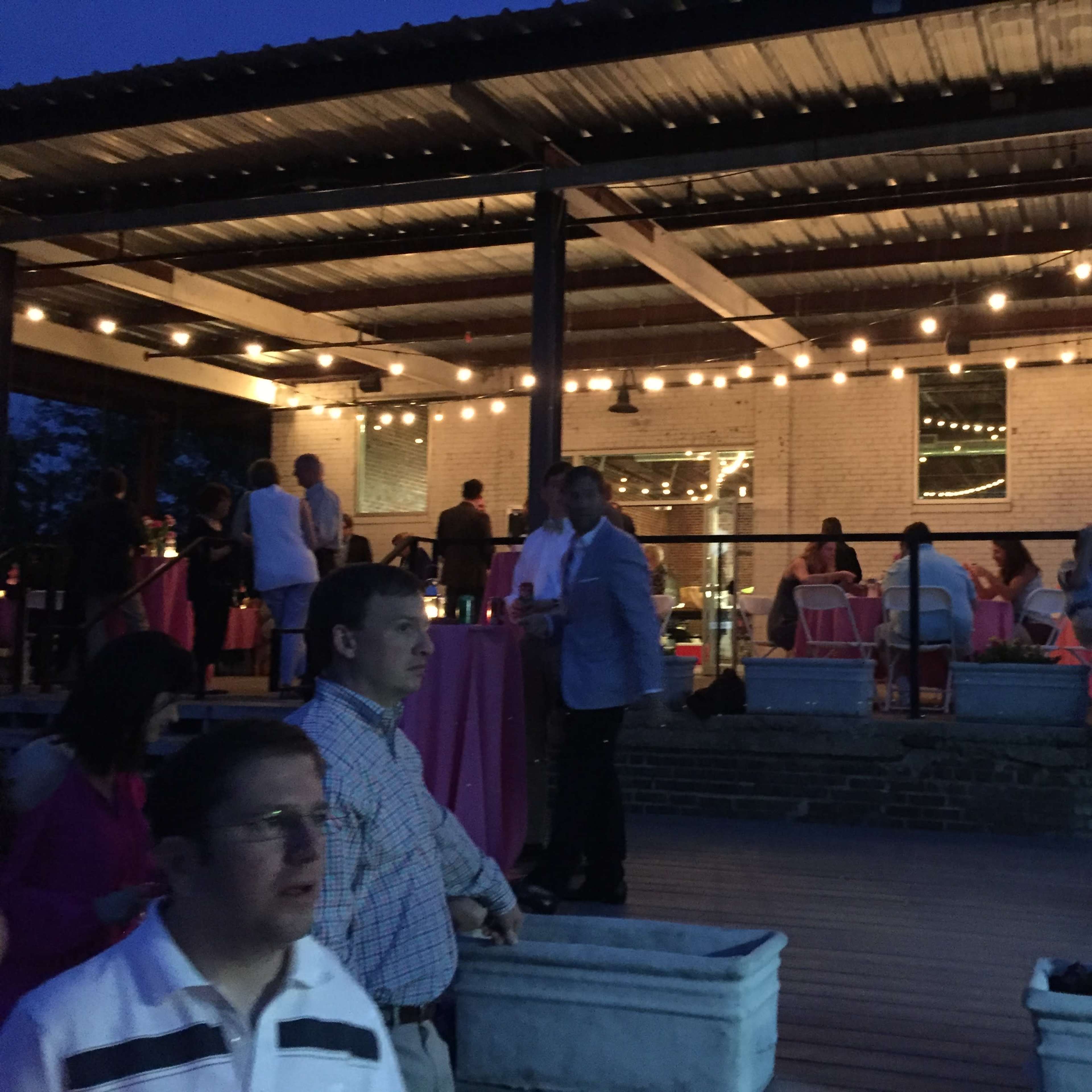 A group of people gathers on an outdoor patio, illuminated by string lights, while dining and socializing during the evening.