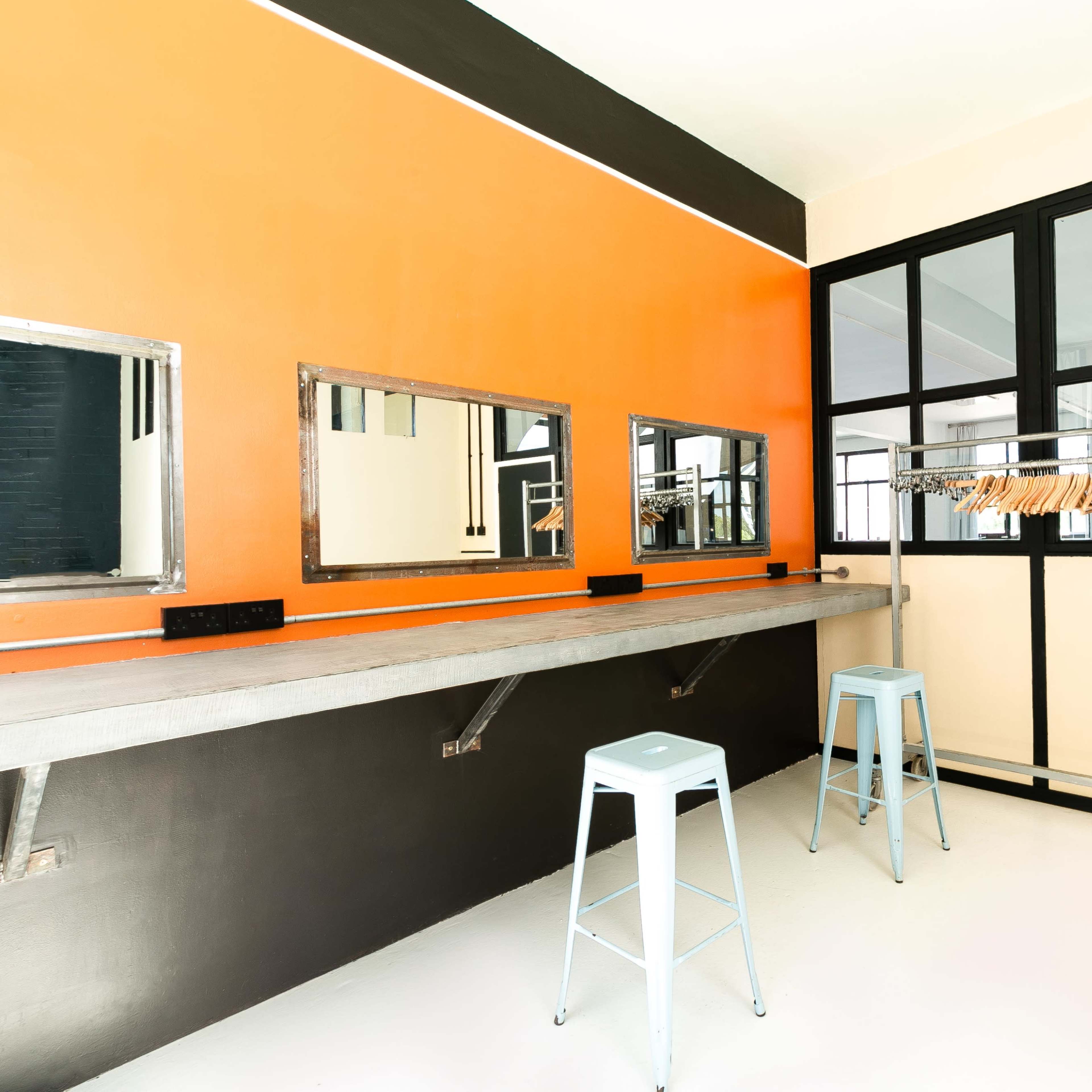A minimalist dressing room with three mirrors, a hanging rack for clothes, and two metal stools against an orange and black wall.