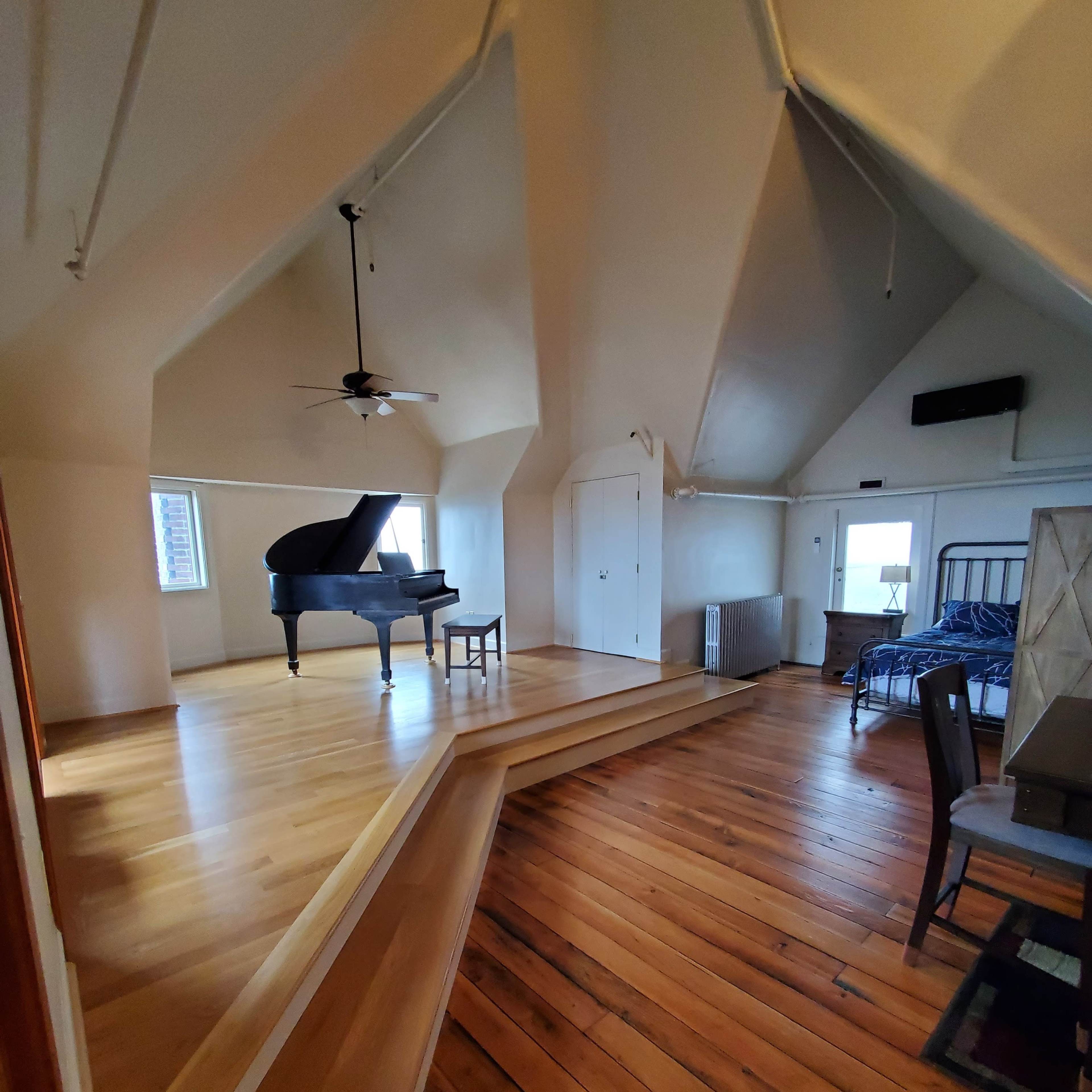 A spacious room with a grand piano on a raised platform, hardwood flooring, and large windows.