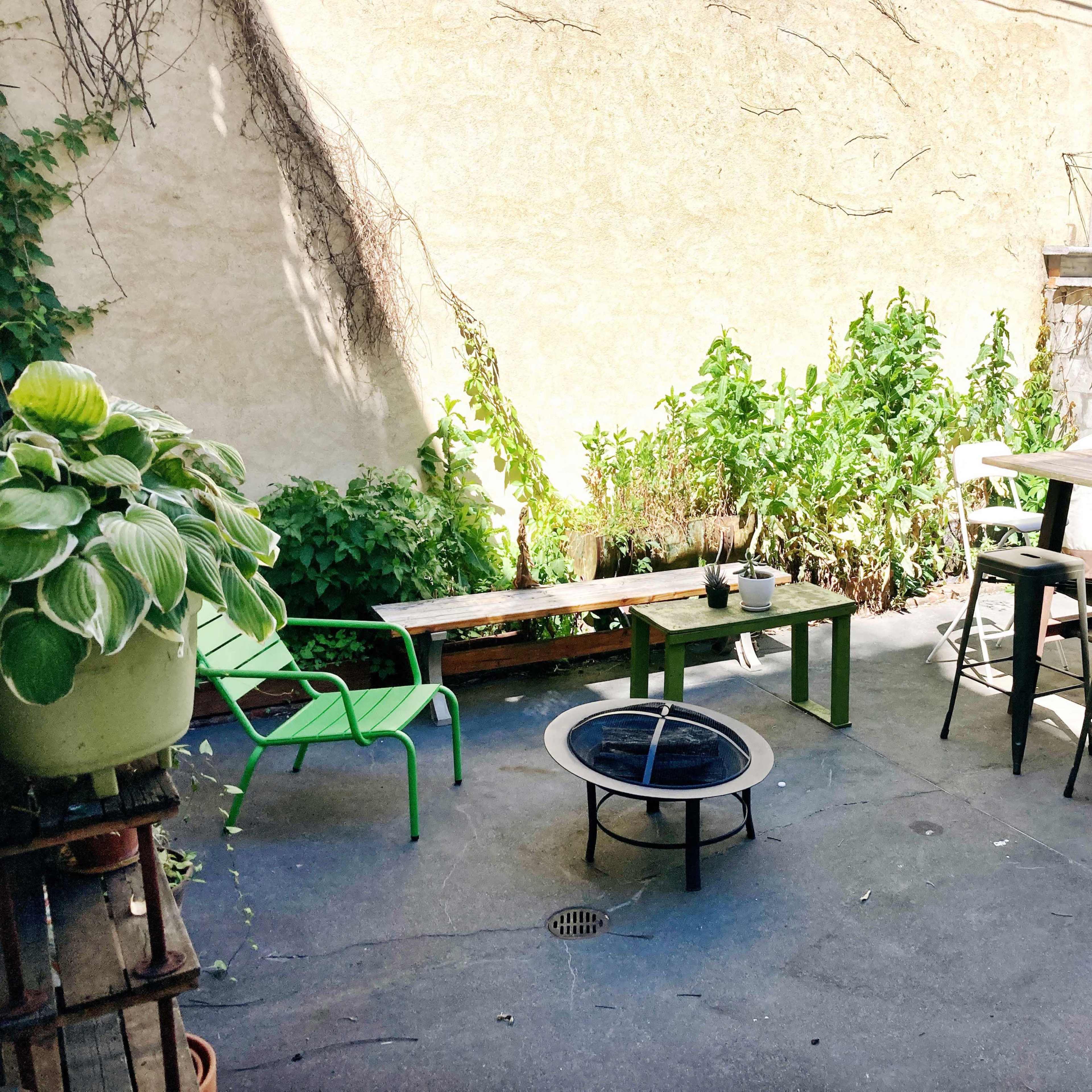 The scene shows a small outdoor patio with green chairs, a wooden table, a fire pit, and surrounding greenery against a bare wall.