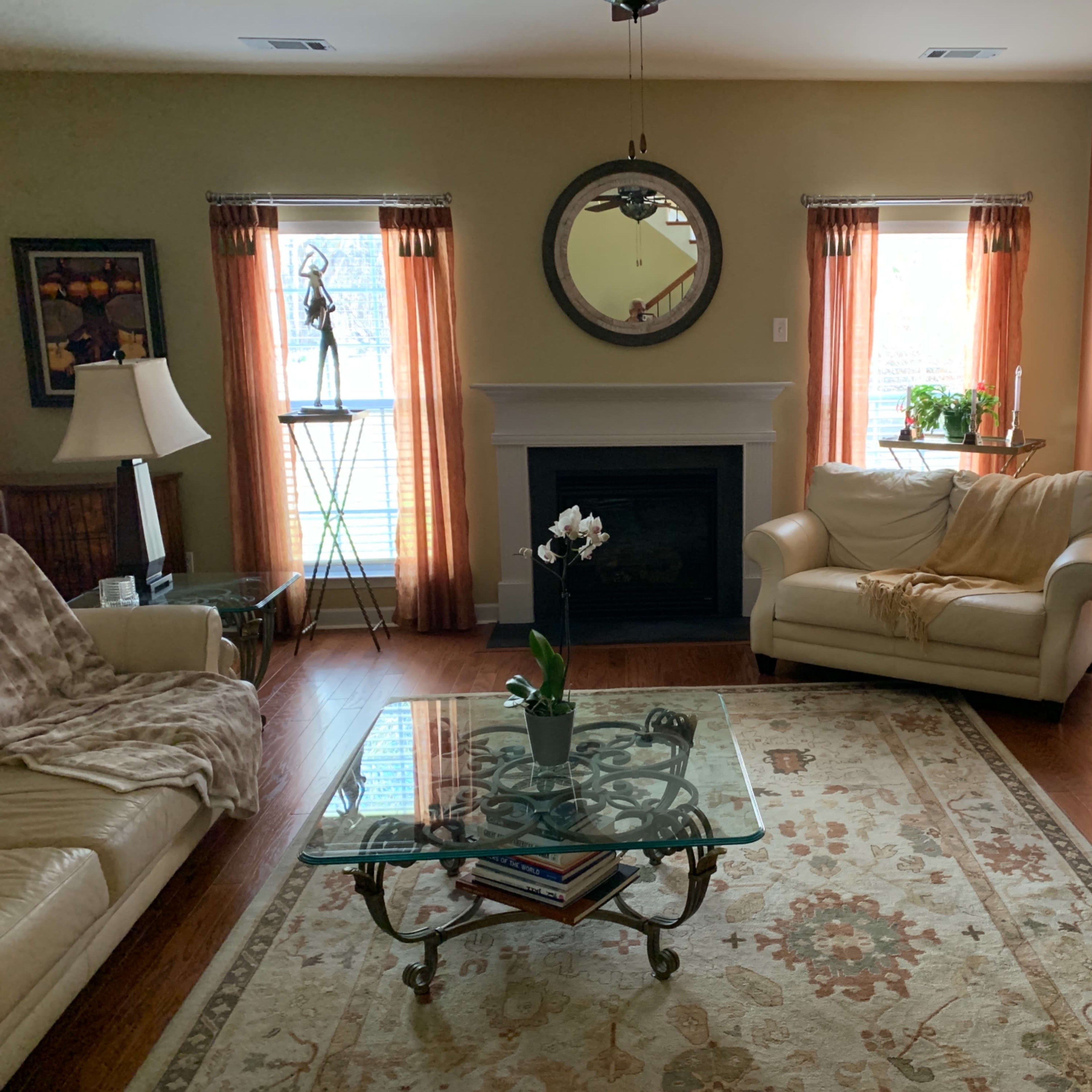 A living room with a beige couch, a glass coffee table on a patterned rug, and windows with orange curtains, all surrounding a fireplace.