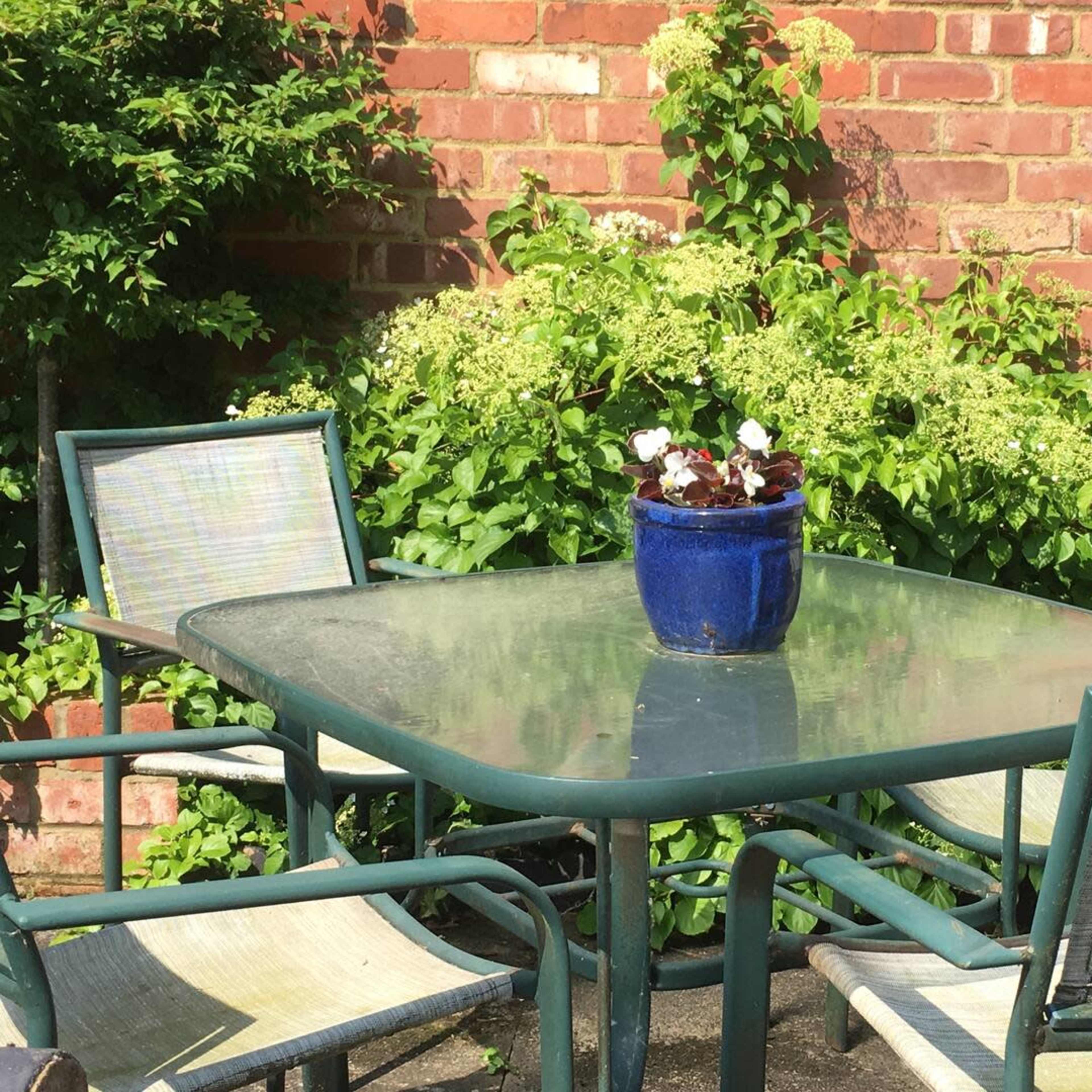 A glass-top table surrounded by four chairs and a blue pot of flowers sits against a brick wall and greenery.