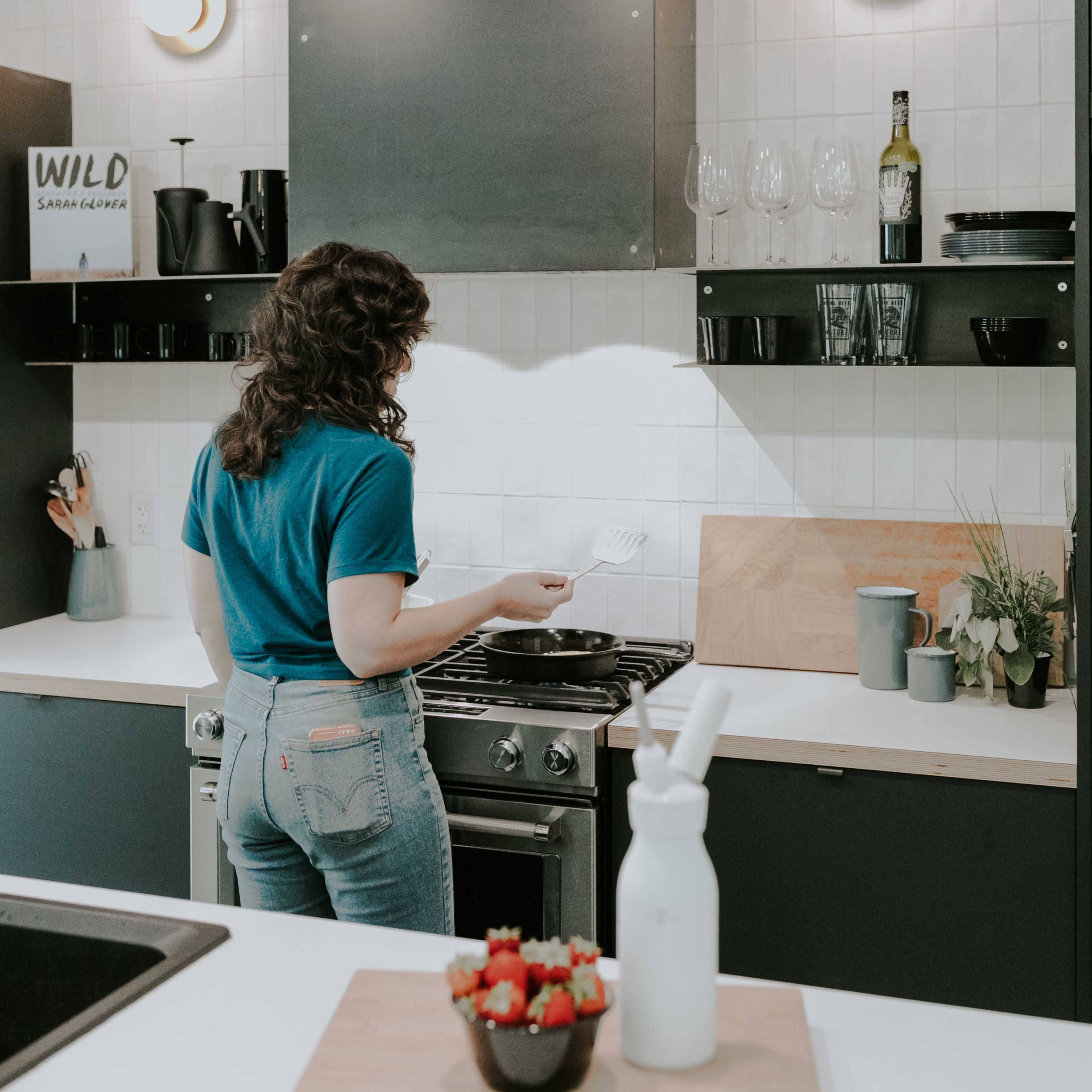 A person with curly hair stands at a stove in a modern kitchen, preparing food.