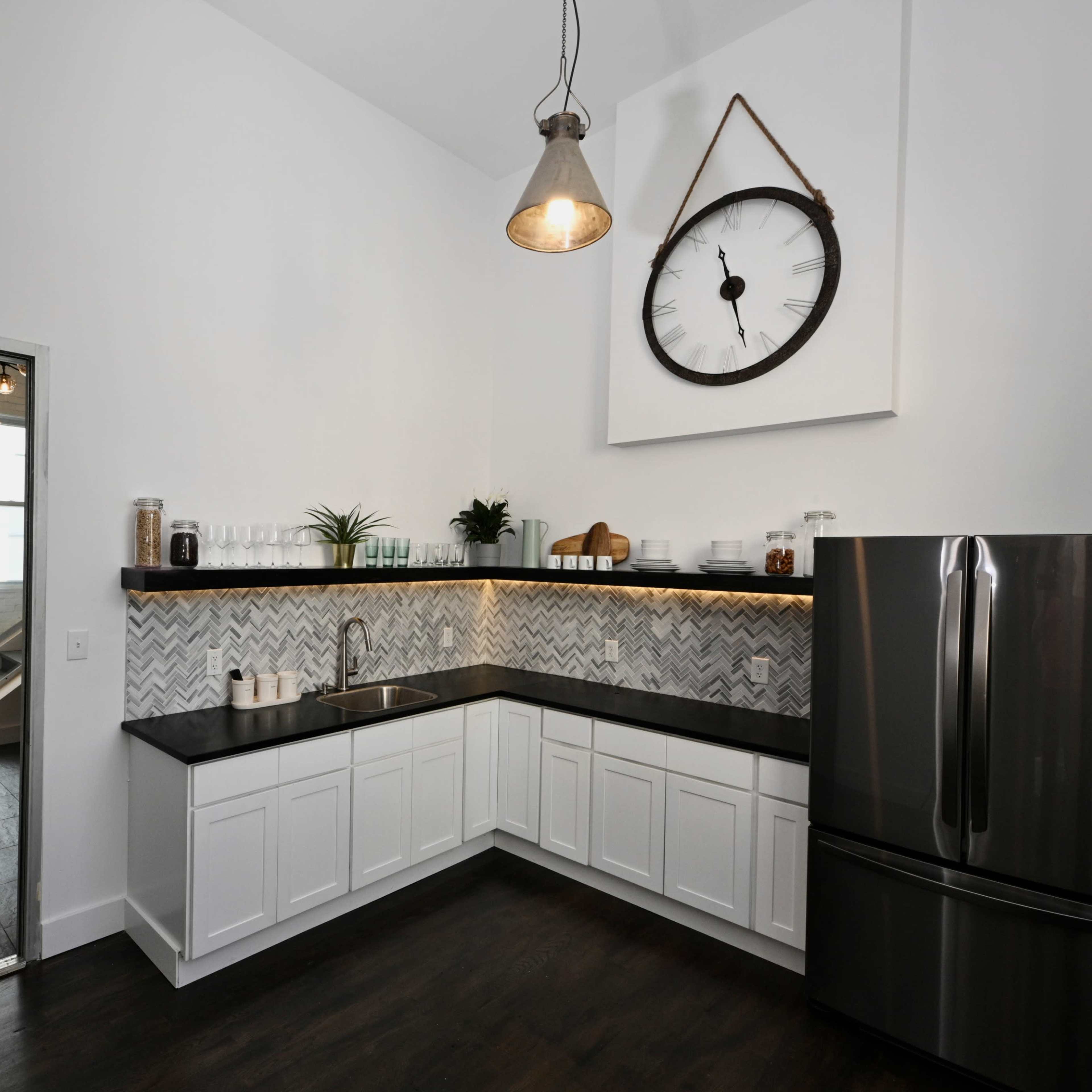 A modern kitchen with white cabinets, a dark countertop, herringbone-style backsplash, and a large wall clock.