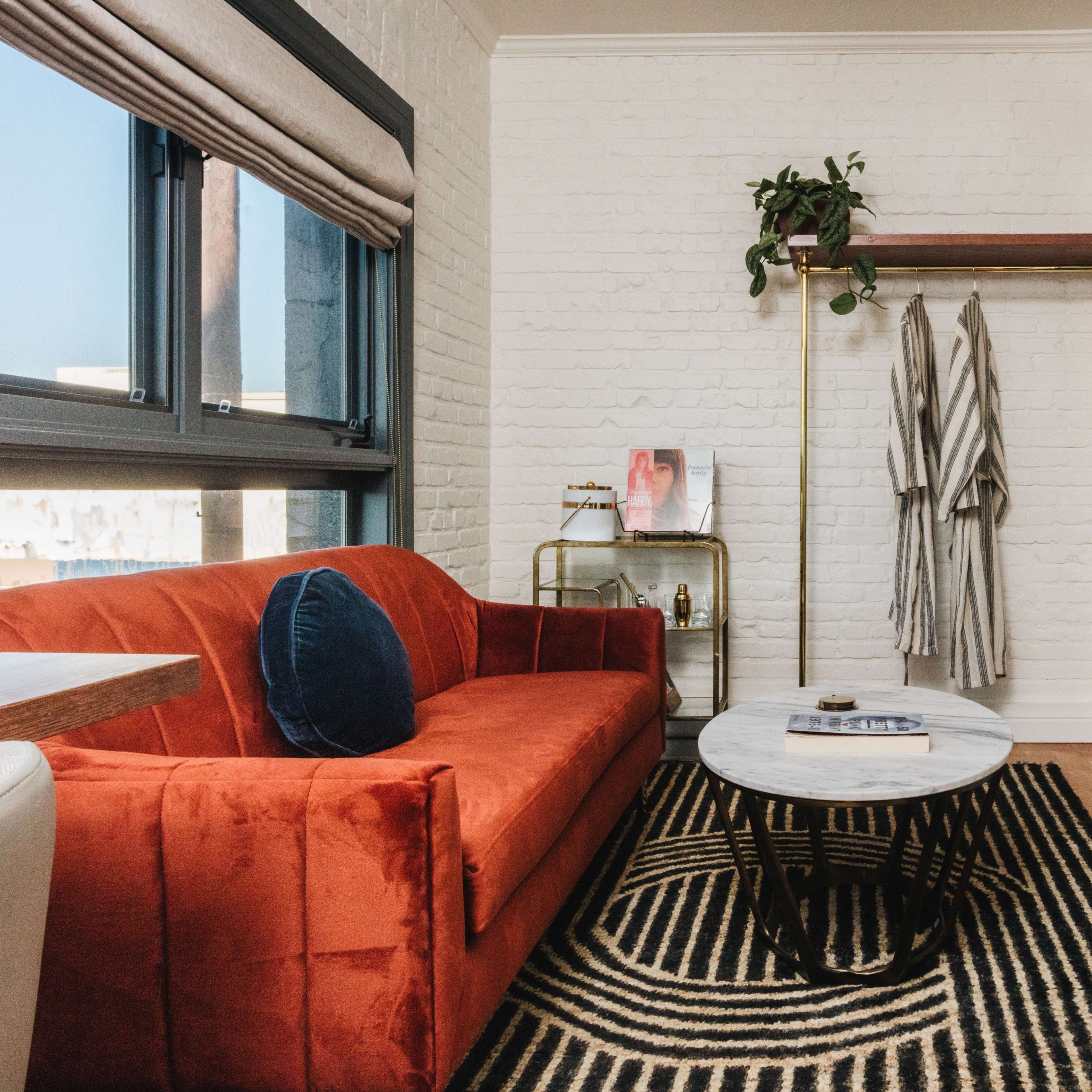 A cozy living space featuring an orange sofa, a round coffee table, a patterned rug, and a clothing rack with hanging robes next to a window.