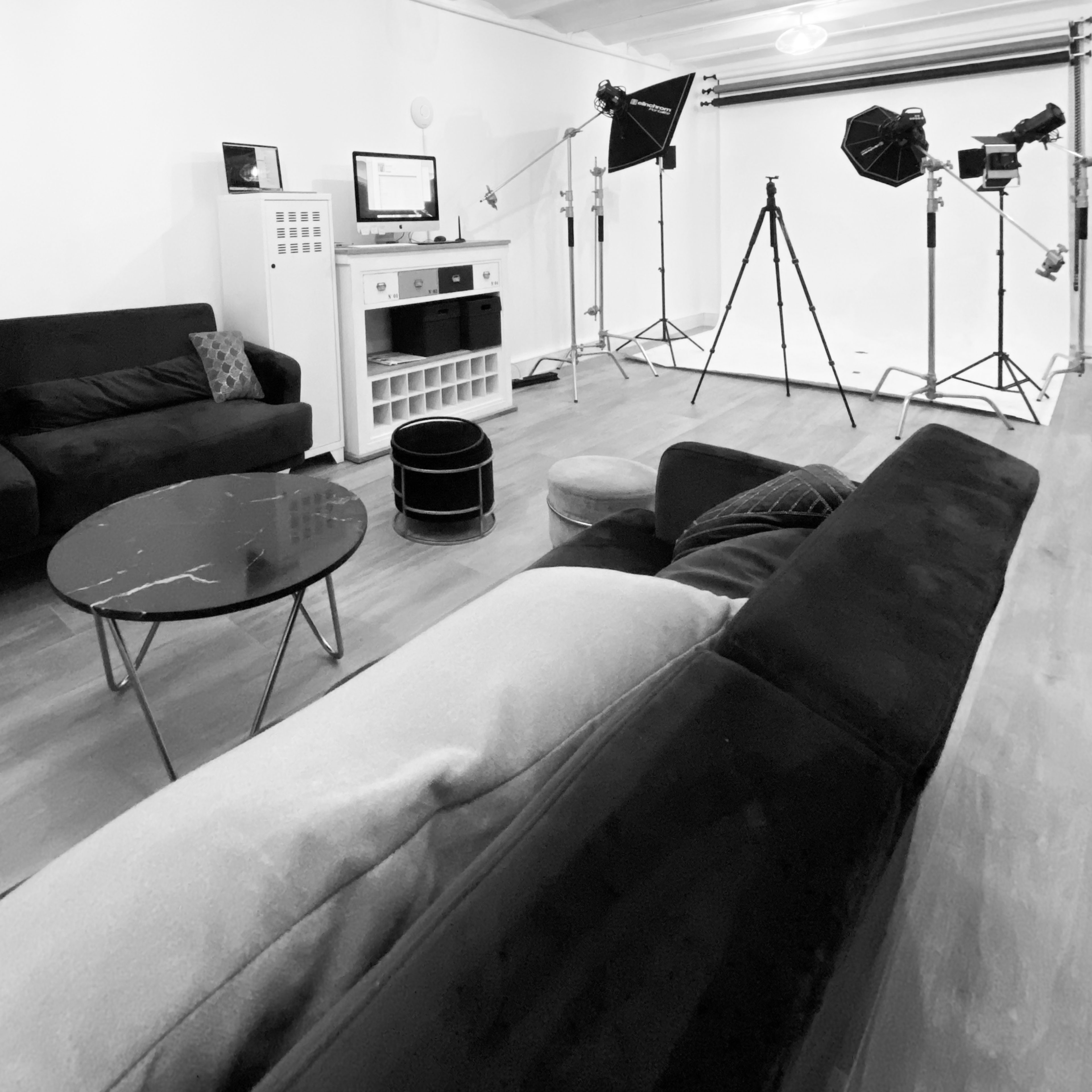 A modern living room setup with black sofas, a coffee table, and studio lighting equipment positioned in the background.