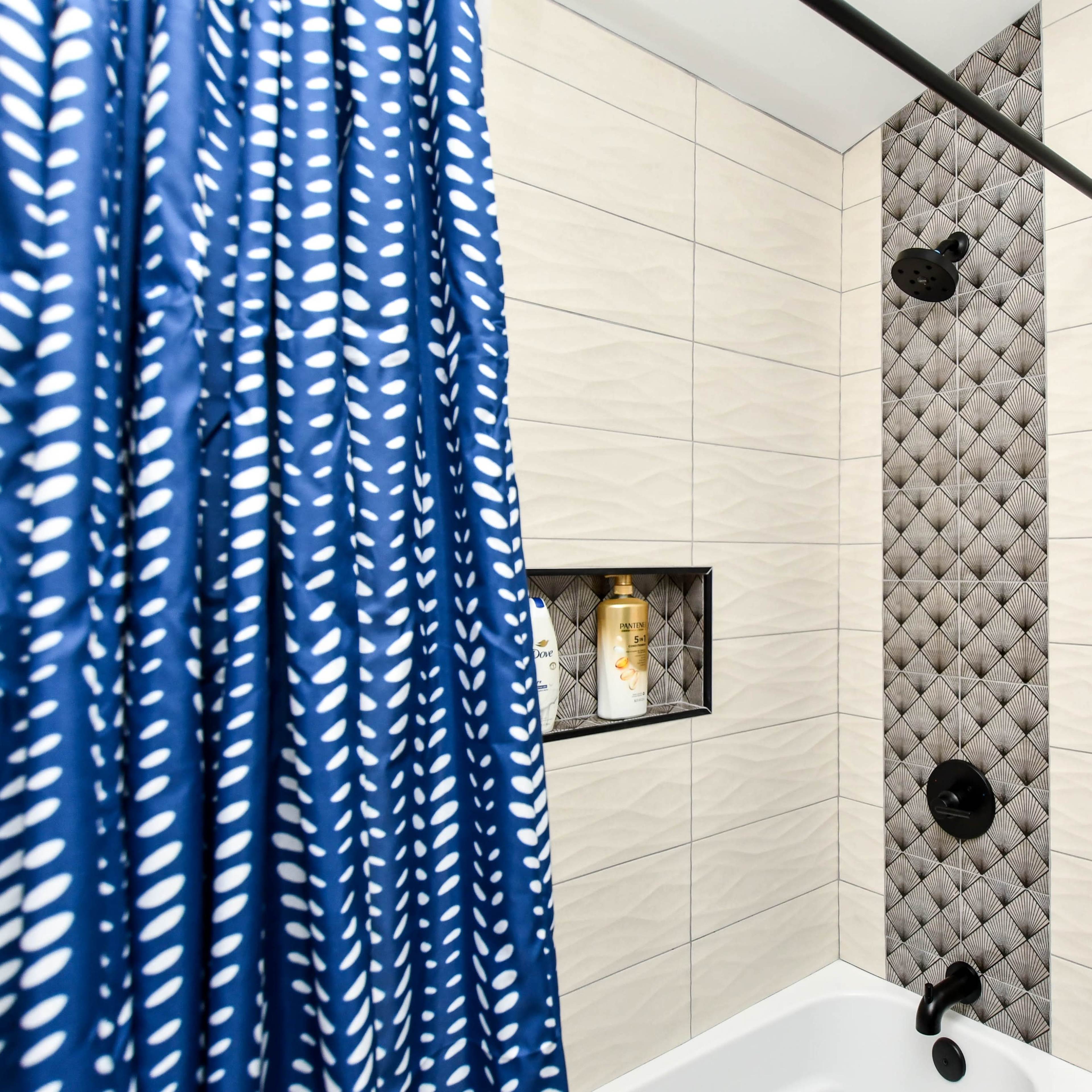 A bathroom shower area featuring a blue patterned curtain, textured wall tiles, and a recessed shelf with a bottle.