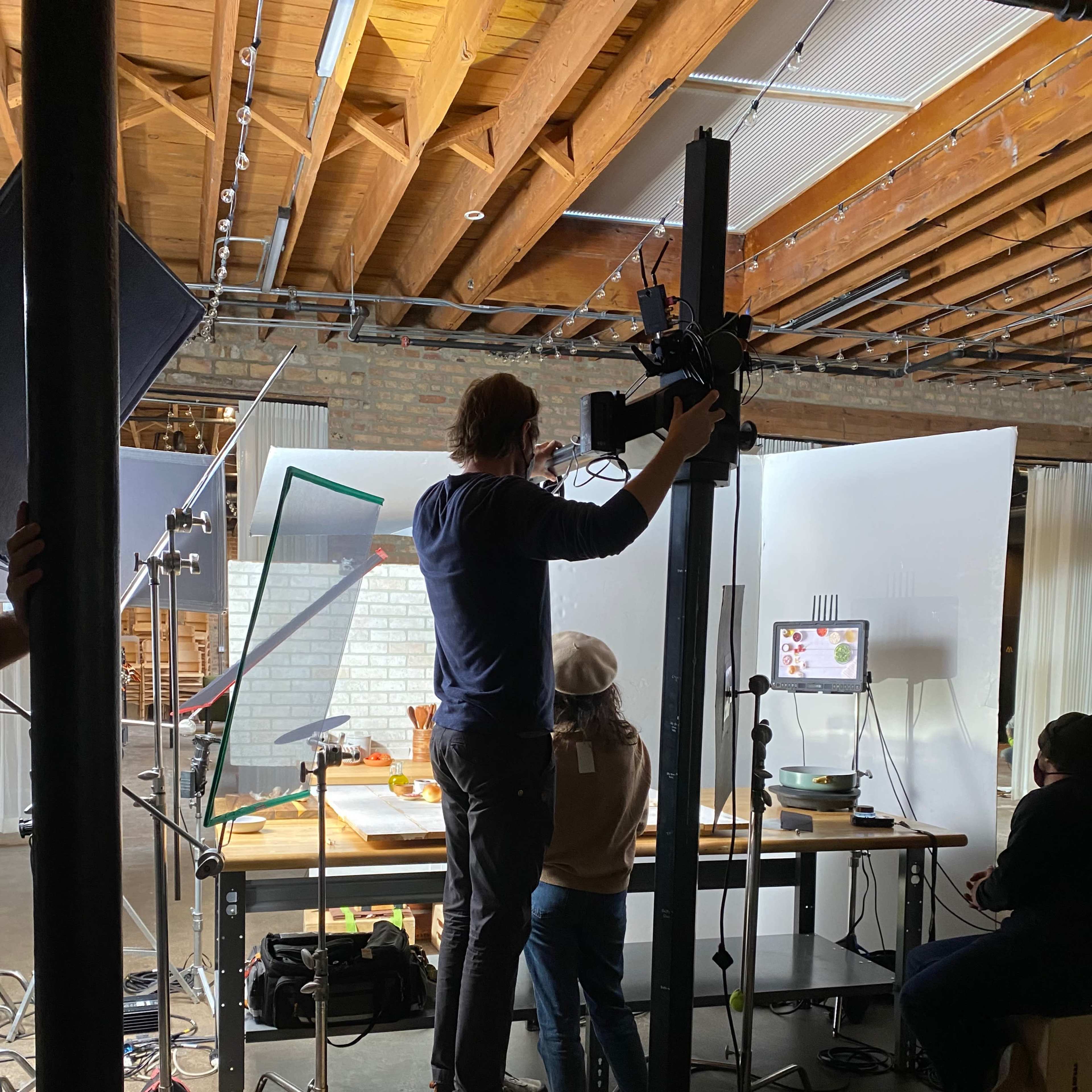 A person is adjusting a camera on a stand while another individual works at a table set up for a photoshoot in a studio.