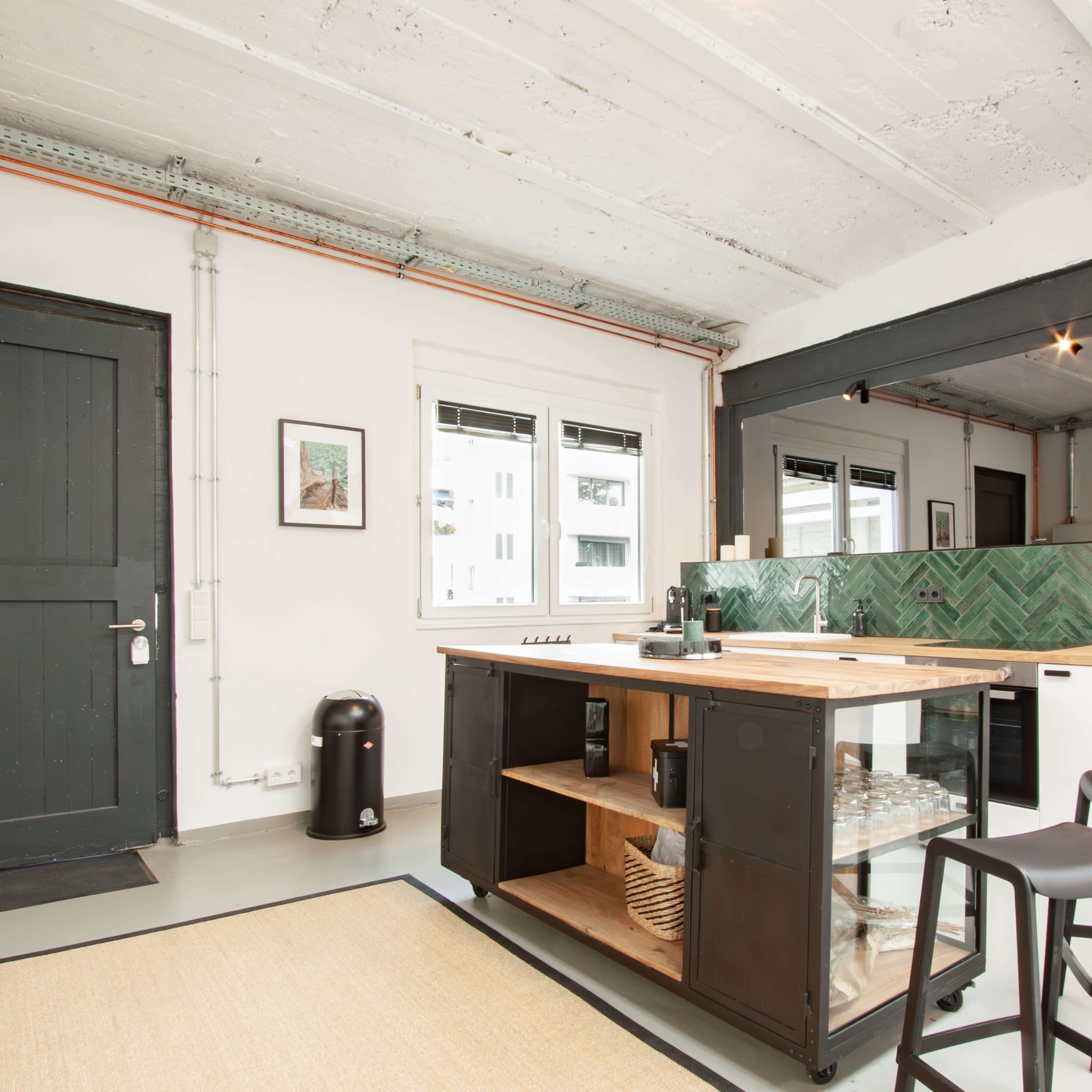 A modern kitchen features a central island with a wooden countertop, black shelves, and a green tile backsplash, alongside a black trash can and a small fridge.