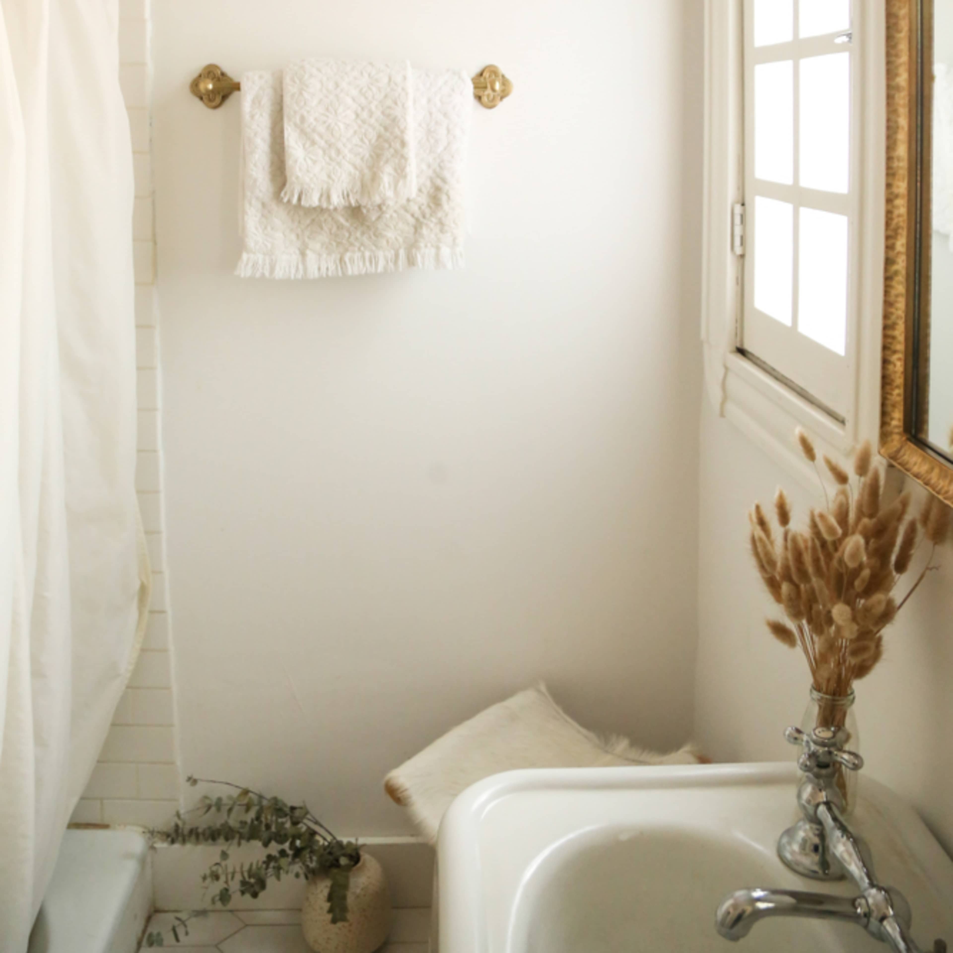 A simplistic bathroom with a bathtub, a shower curtain, a towel rack holding a white towel, and a mirror beside a decorative vase with dried plants.