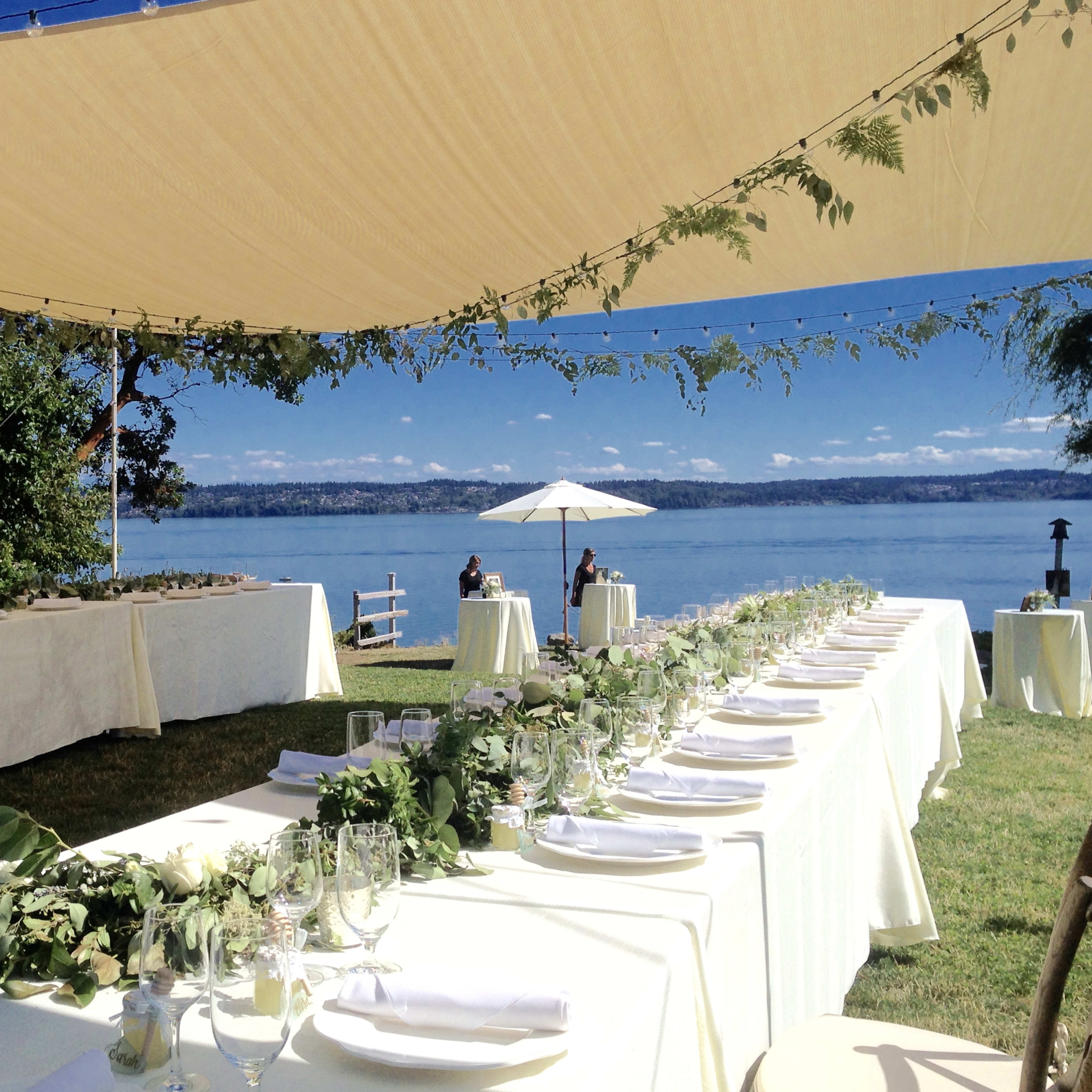 A long, elegantly set dining table is arranged under a shaded canopy beside a calm waterfront, with greenery and white tableware.