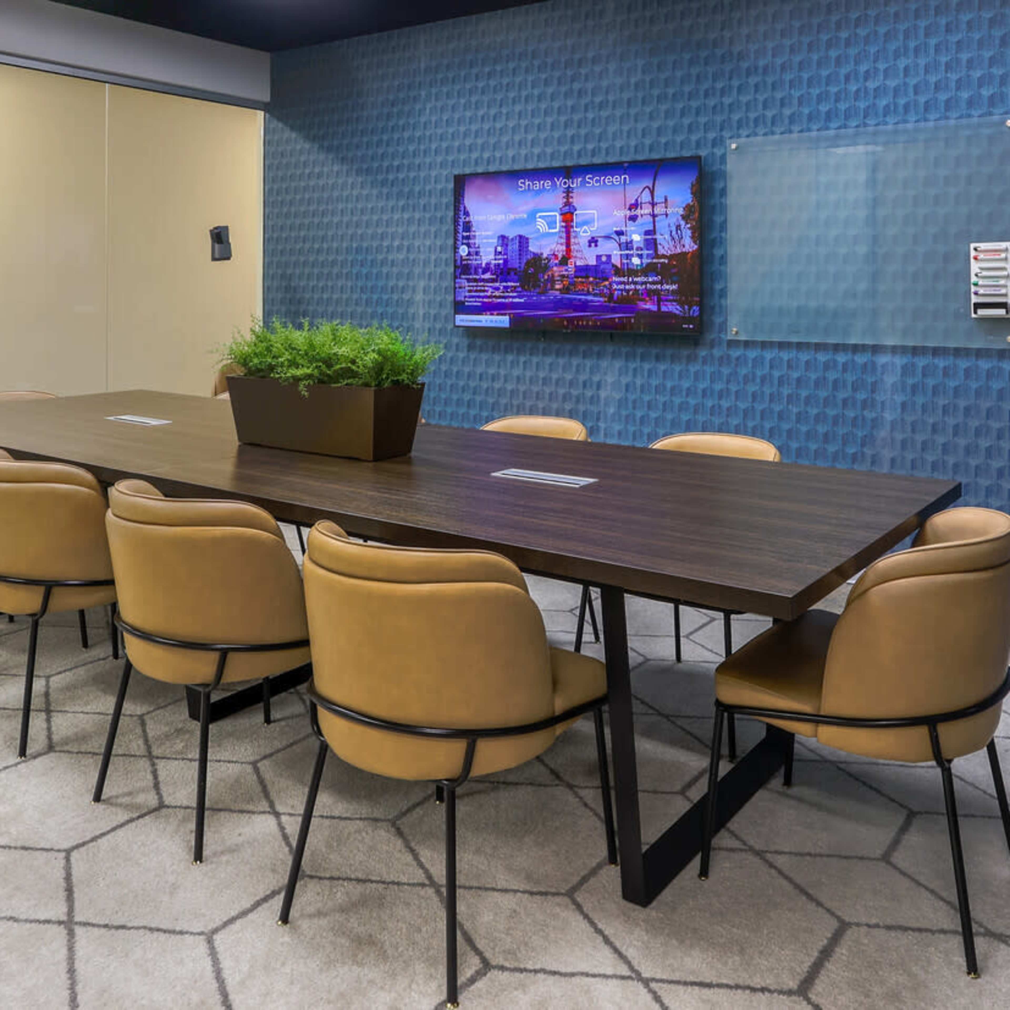 A modern conference room with a long wooden table surrounded by eight brown chairs, a television mounted on the wall, and a potted plant as a centerpiece.