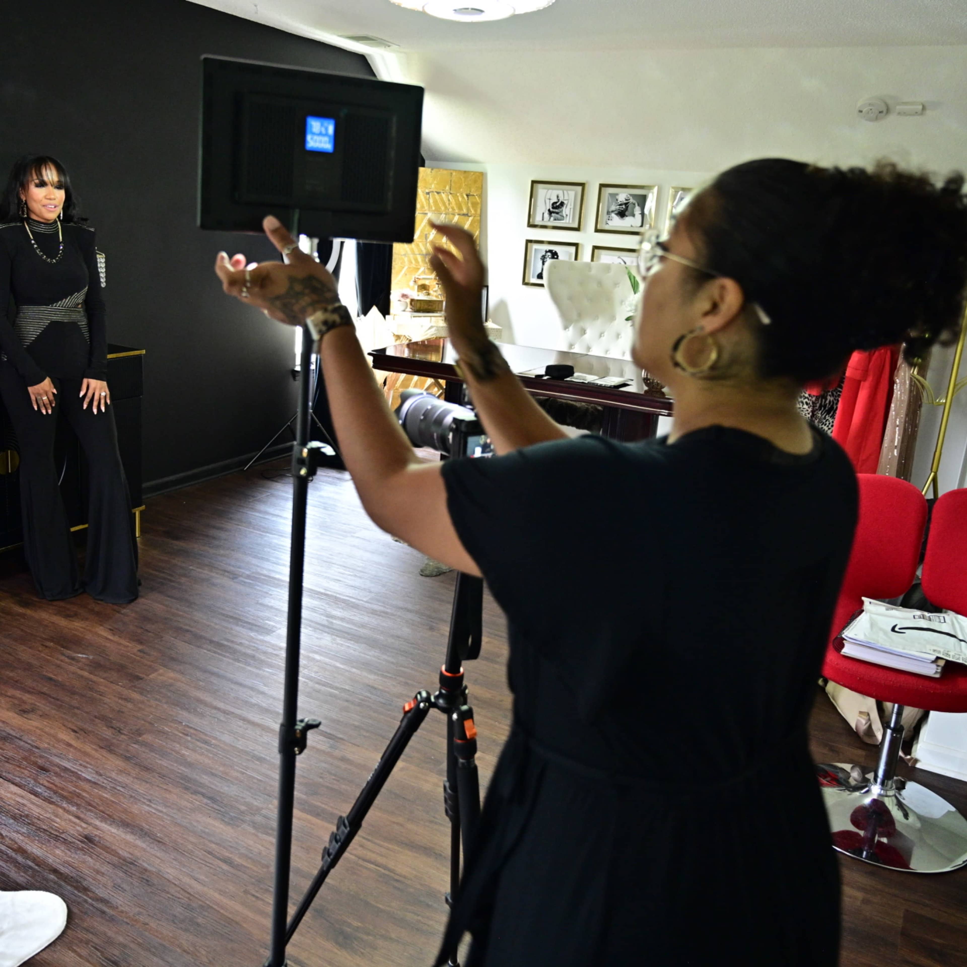 A woman in a black outfit poses for a photo shoot in a stylishly decorated room while another woman adjusts the camera on a tripod.