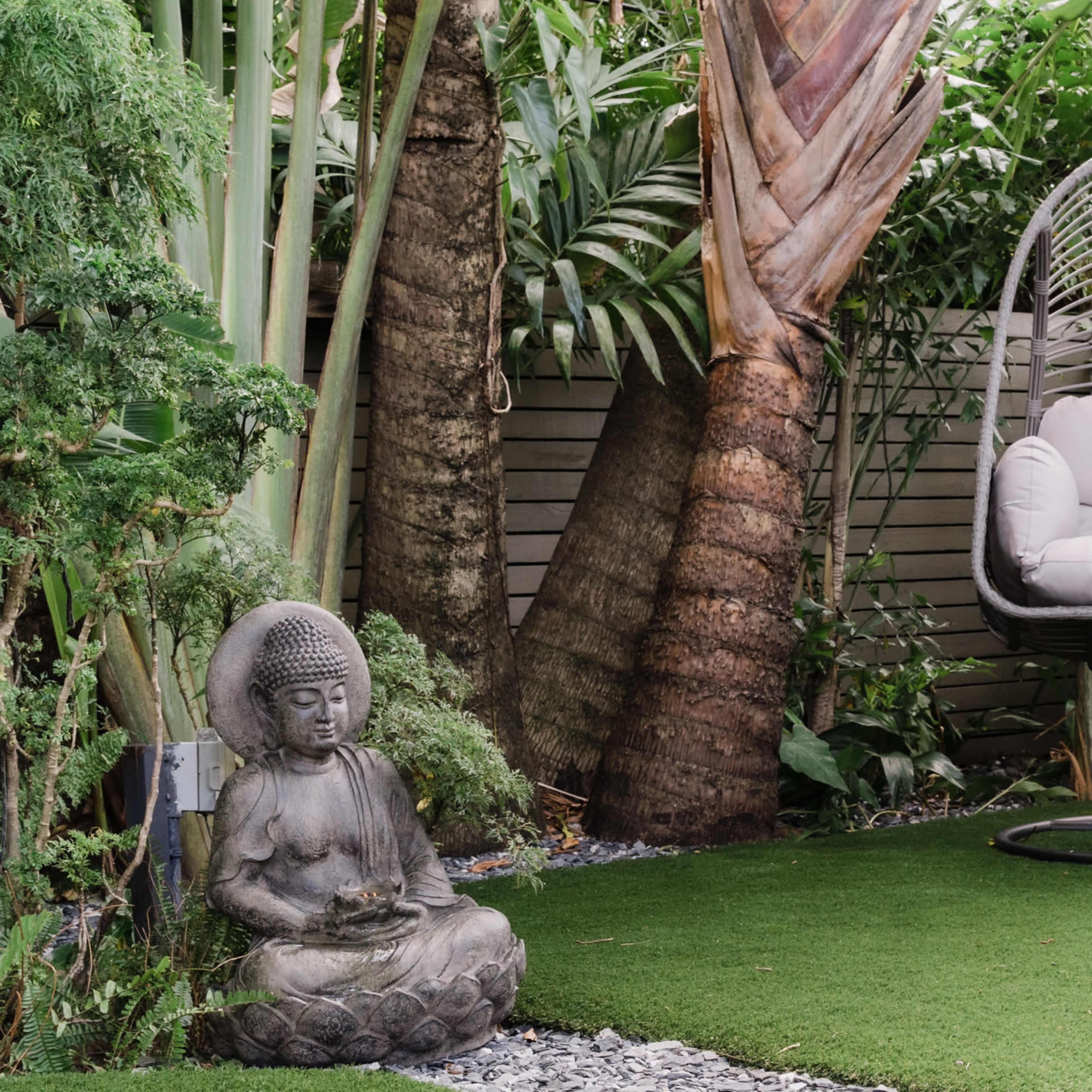 A serene garden scene with a hanging chair and a stone Buddha statue surrounded by lush greenery and artificial grass.