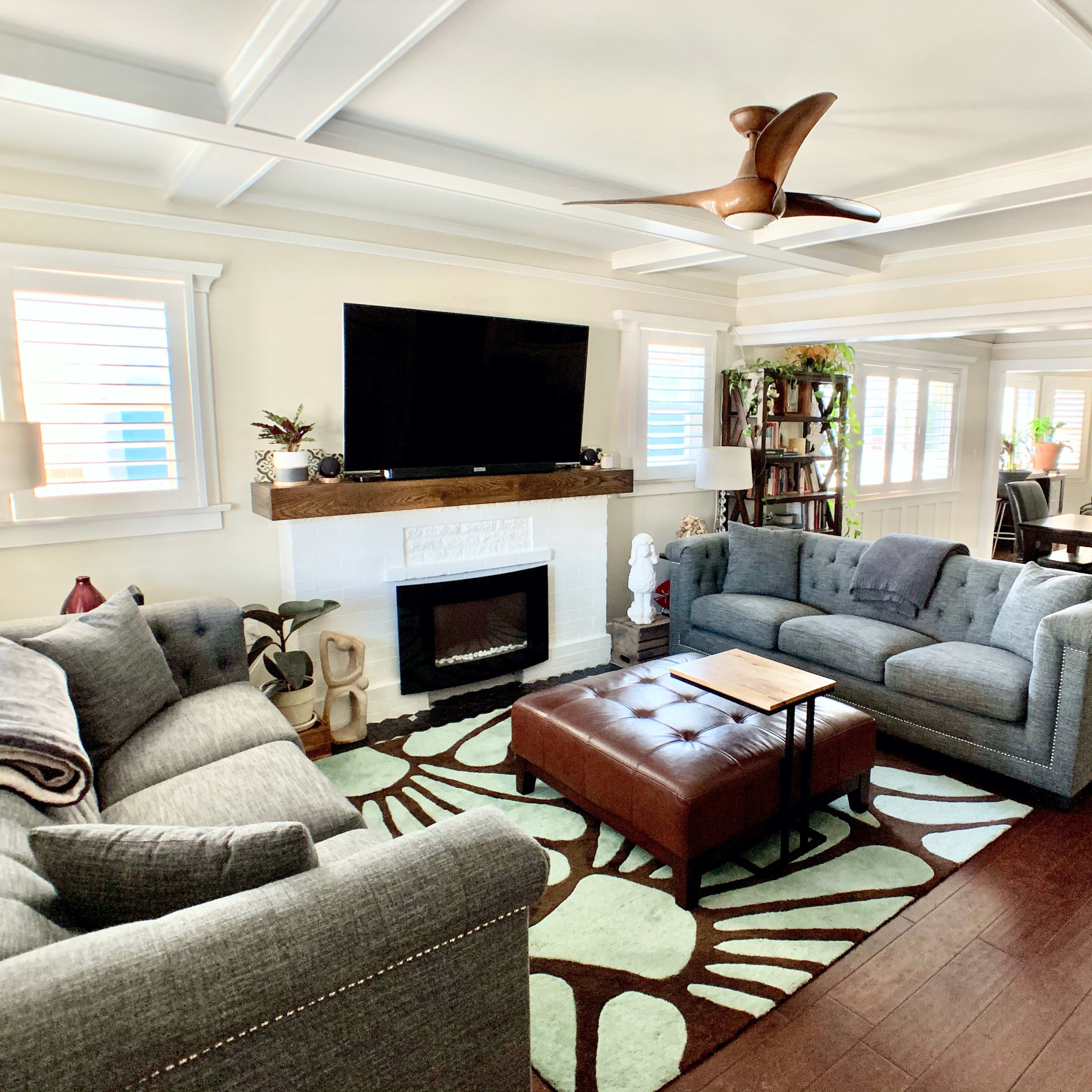 The living room features two gray sofas facing a coffee table, with a flat-screen TV mounted above a white fireplace, and a dining area visible in the background.