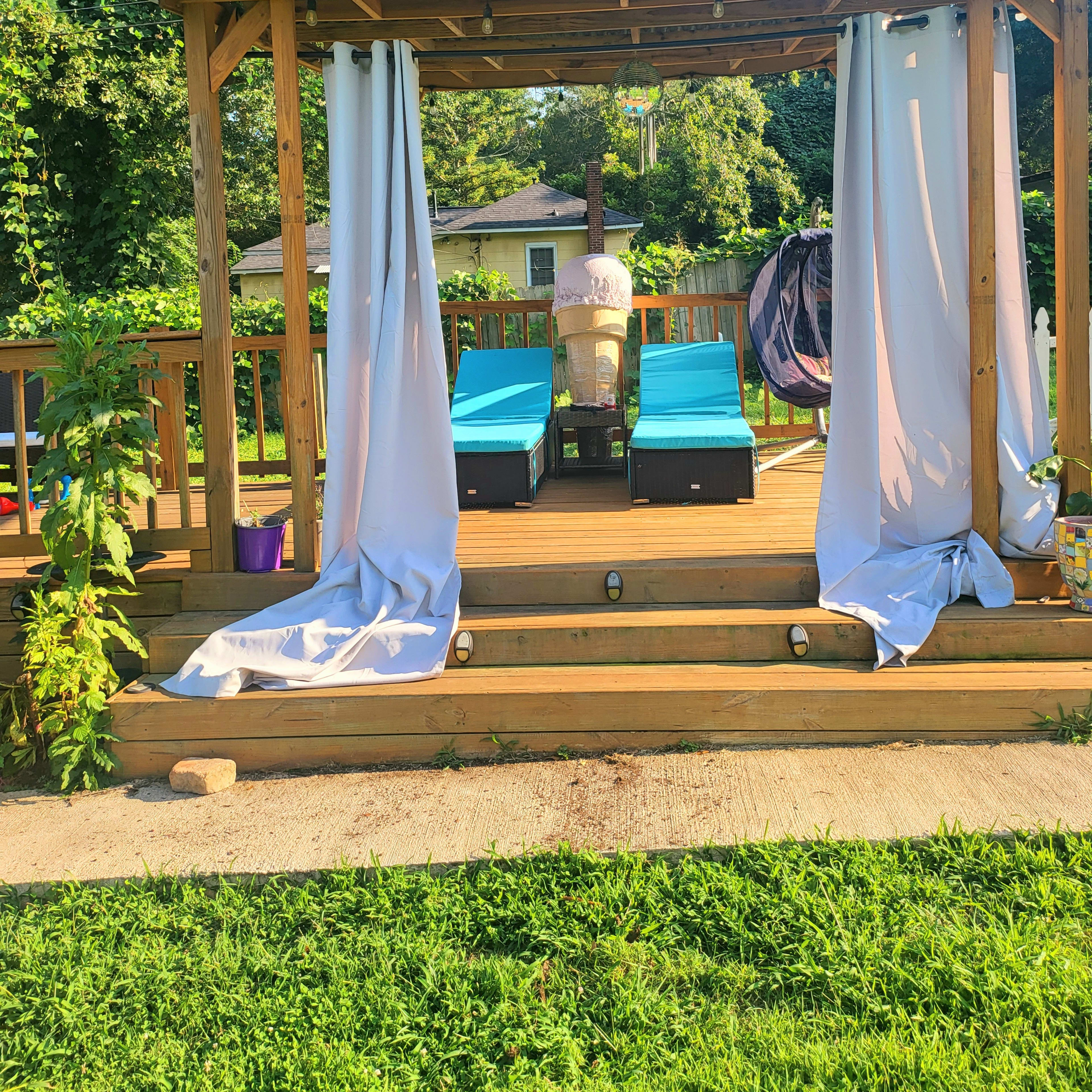 A wooden patio with two blue lounge chairs and white curtains, surrounded by greenery and a hanging seat.