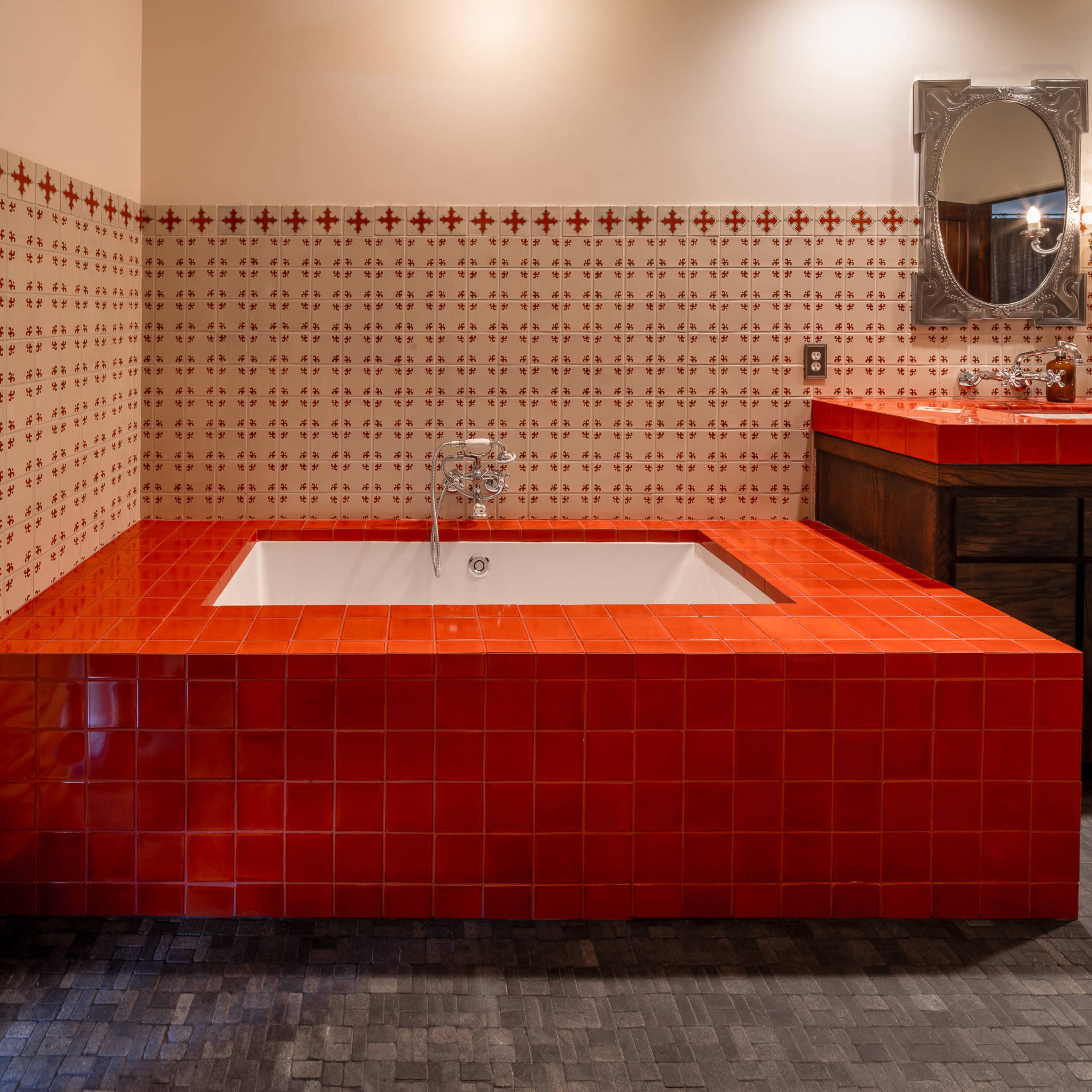 A bathroom featuring a red tiled bathtub, surrounded by patterned wall tiles and a wooden vanity with a mirror above it.