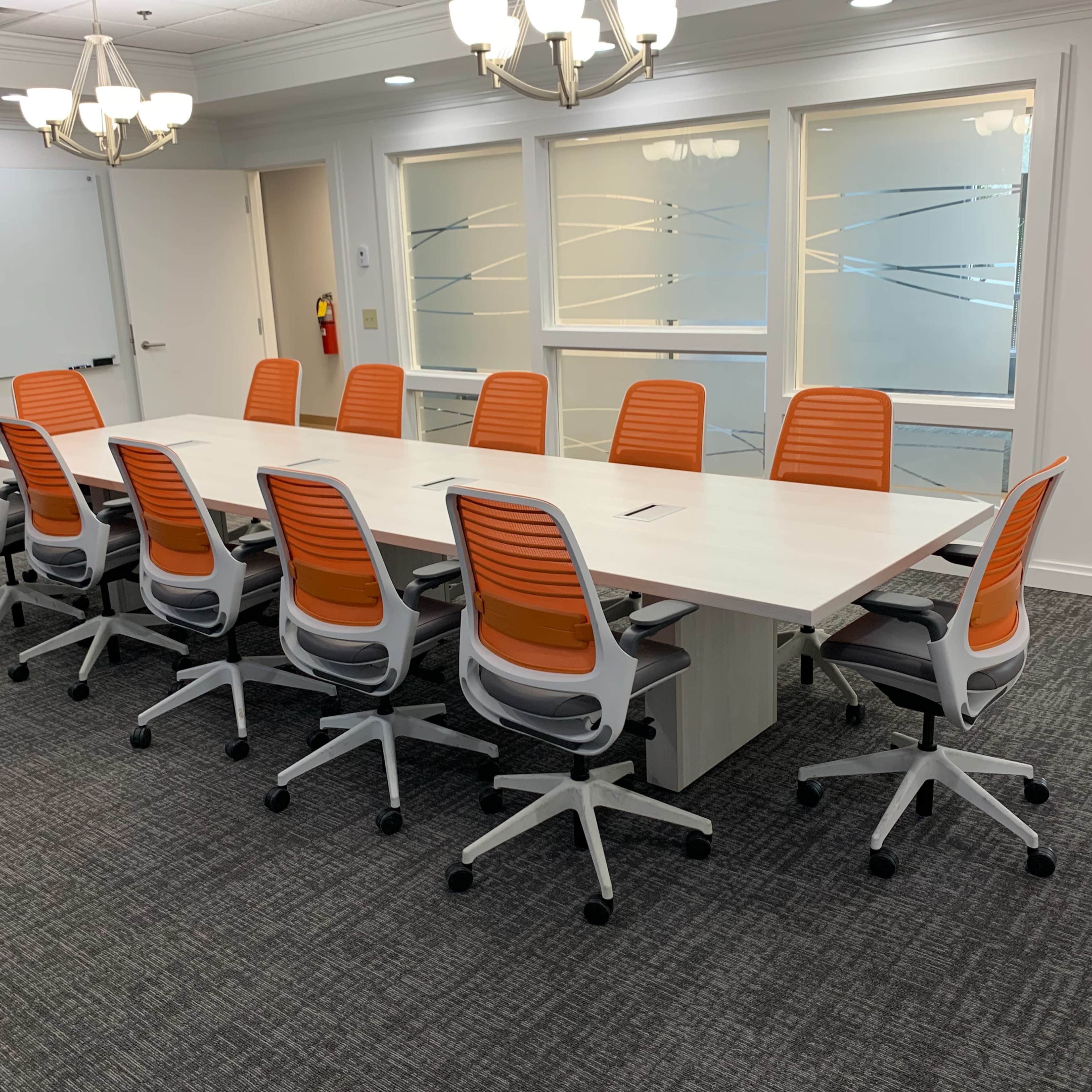 A conference room is set with a long white table surrounded by twelve orange and gray office chairs.