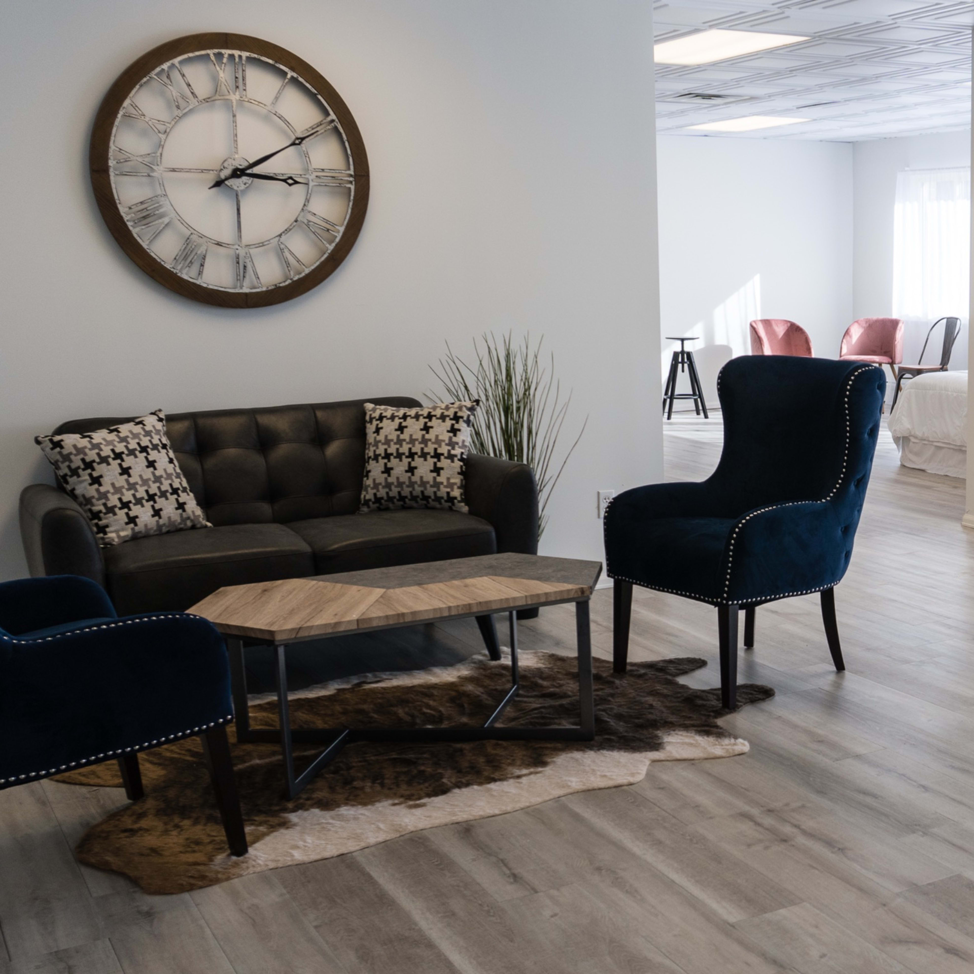 A spacious, well-lit room features a seating area with two blue armchairs and a sofa, accompanied by a wooden coffee table on a rug, while a bright, empty area is visible in the background.