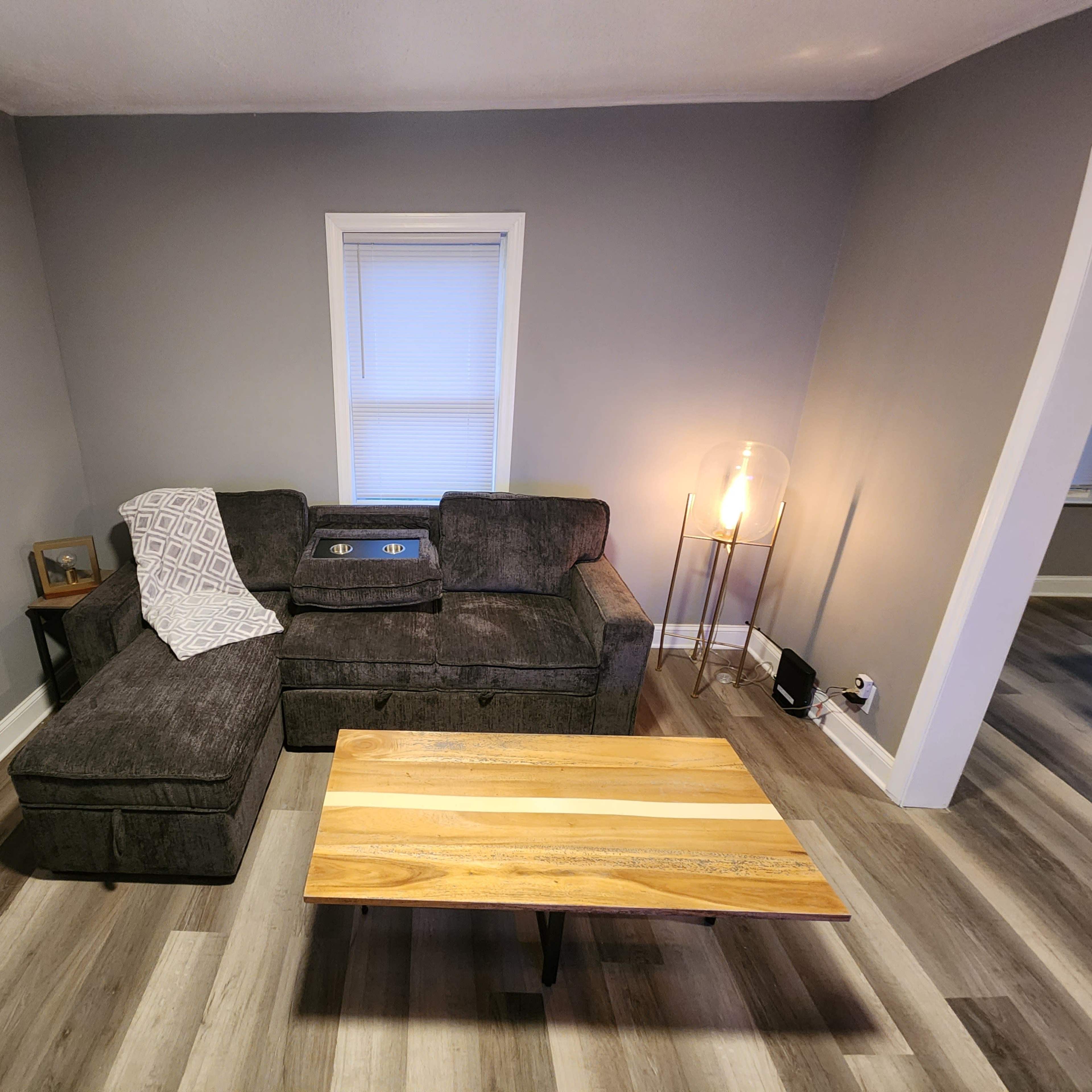 A living room with a dark gray sectional sofa, a wooden coffee table, and a round lamp against a gray wall, with two windows providing natural light.
