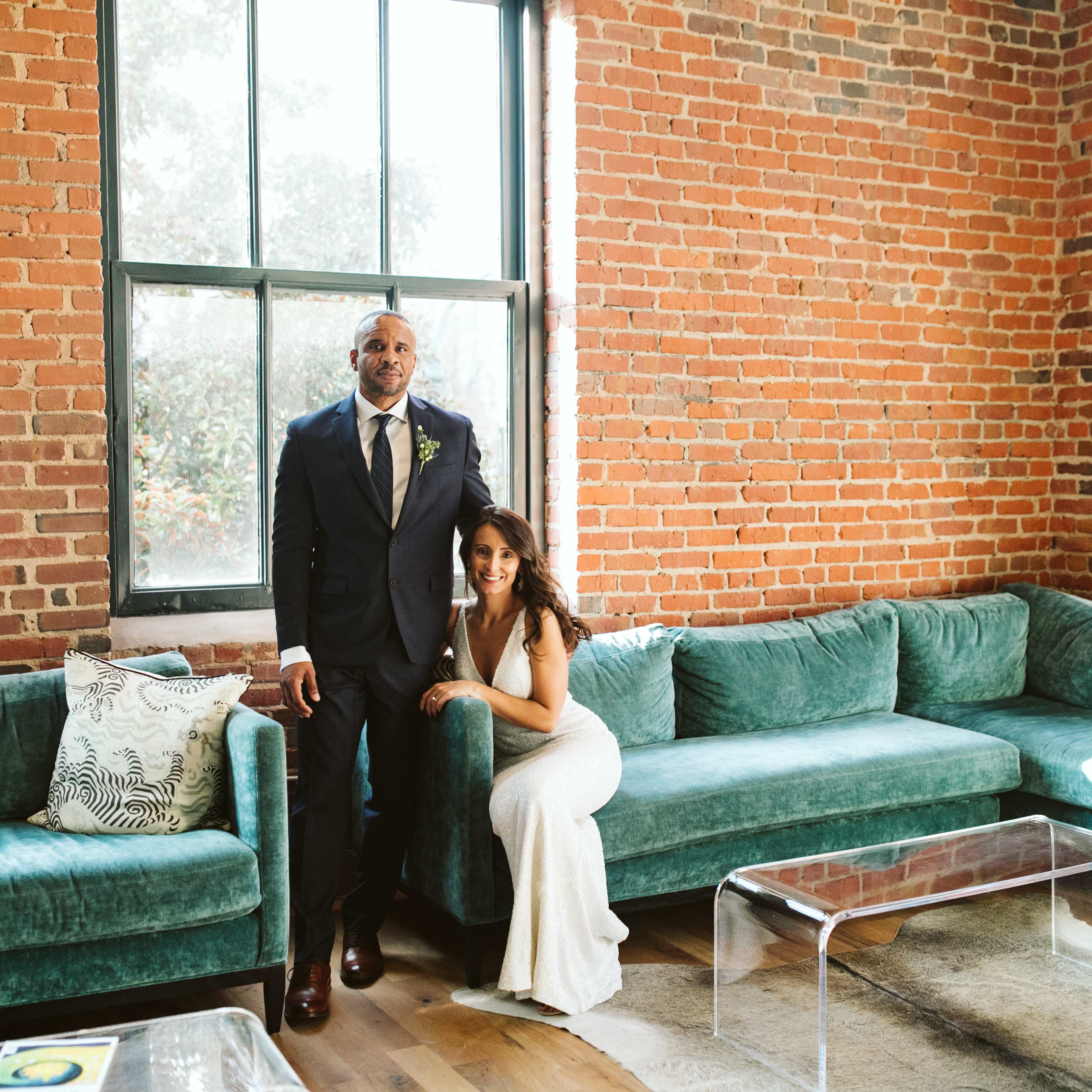 A man in a suit stands beside a woman in a white dress in a room with exposed brick walls and teal sofas.