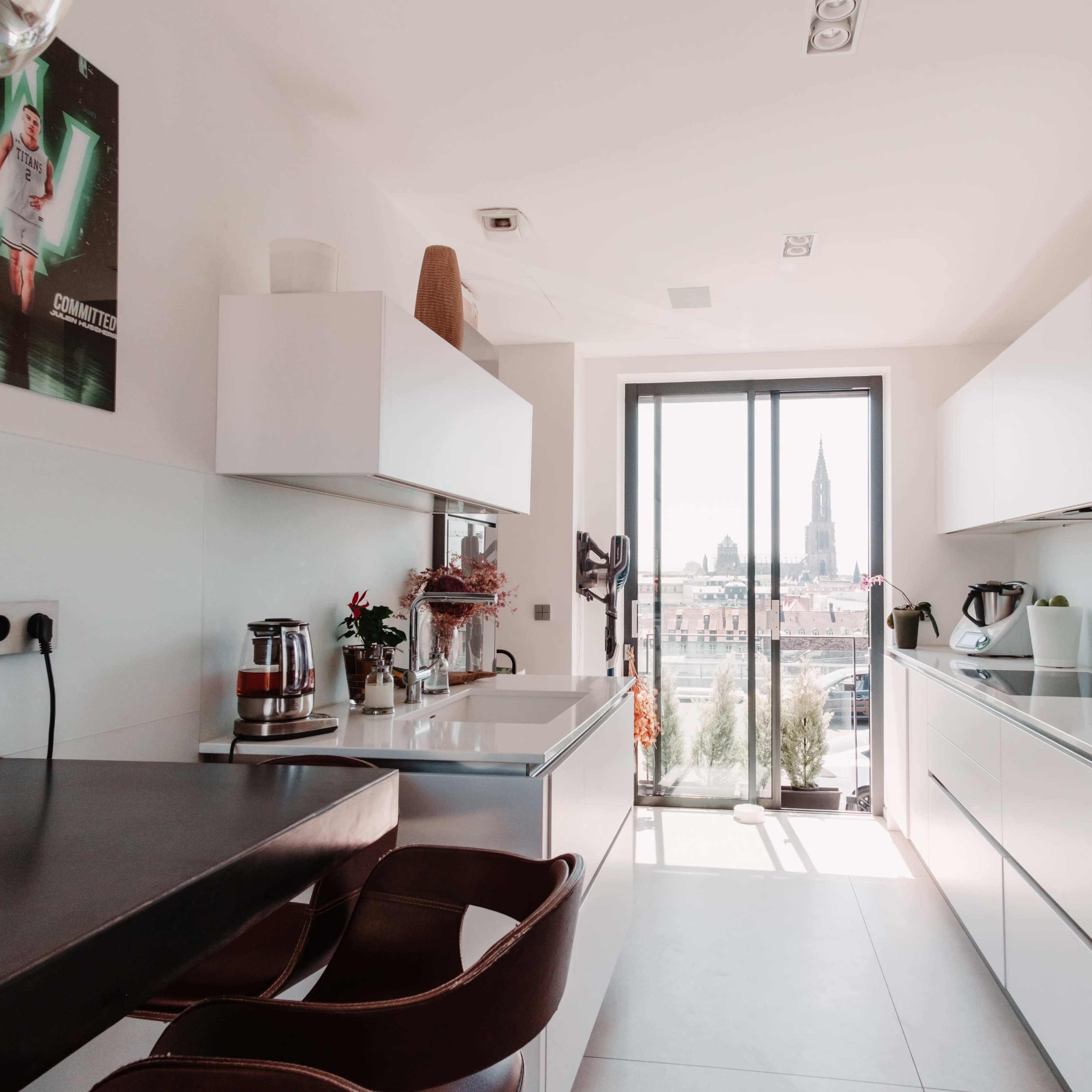 A modern kitchen features sleek white cabinetry, a small breakfast bar with two brown chairs, and a large window showcasing a cityscape with a prominent tower.