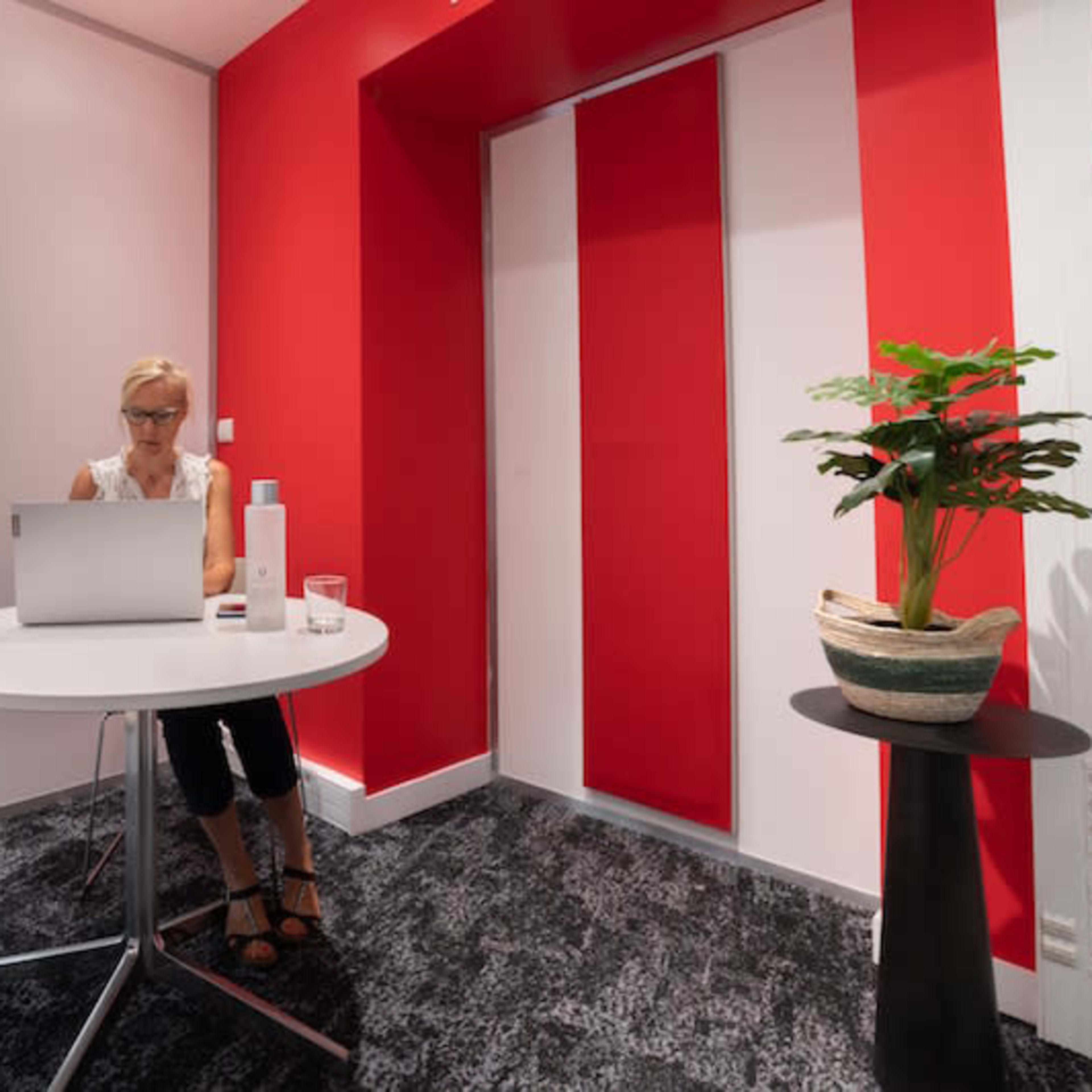 A woman is sitting at a round table with a laptop in a modern office space featuring red and white walls and a potted plant nearby.