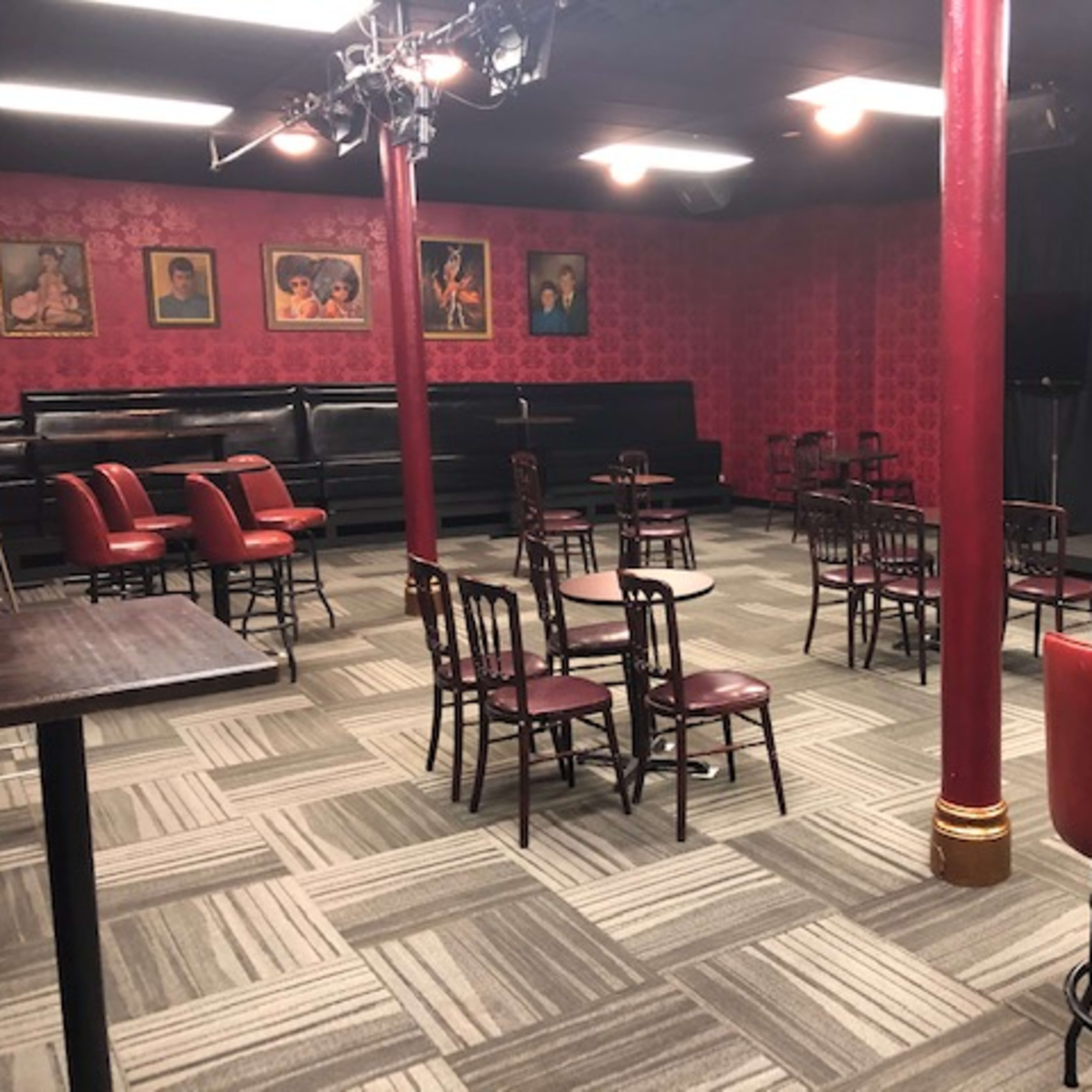 A dimly lit comedy club interior with red walls, empty tables and chairs, and a small stage at the back.