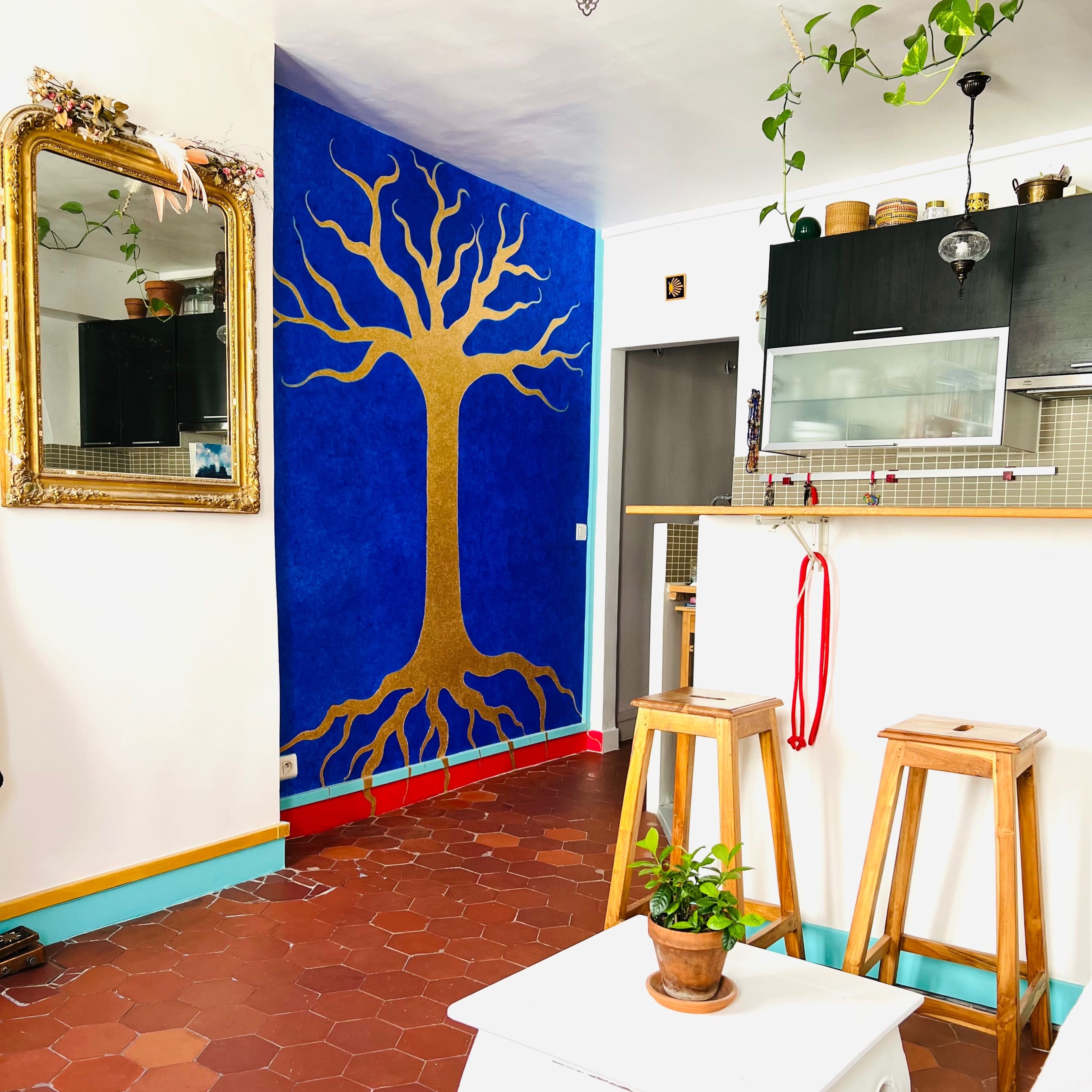 A brightly colored interior space featuring a blue wall with a golden tree design, wooden bar stools, and a decorative mirror.