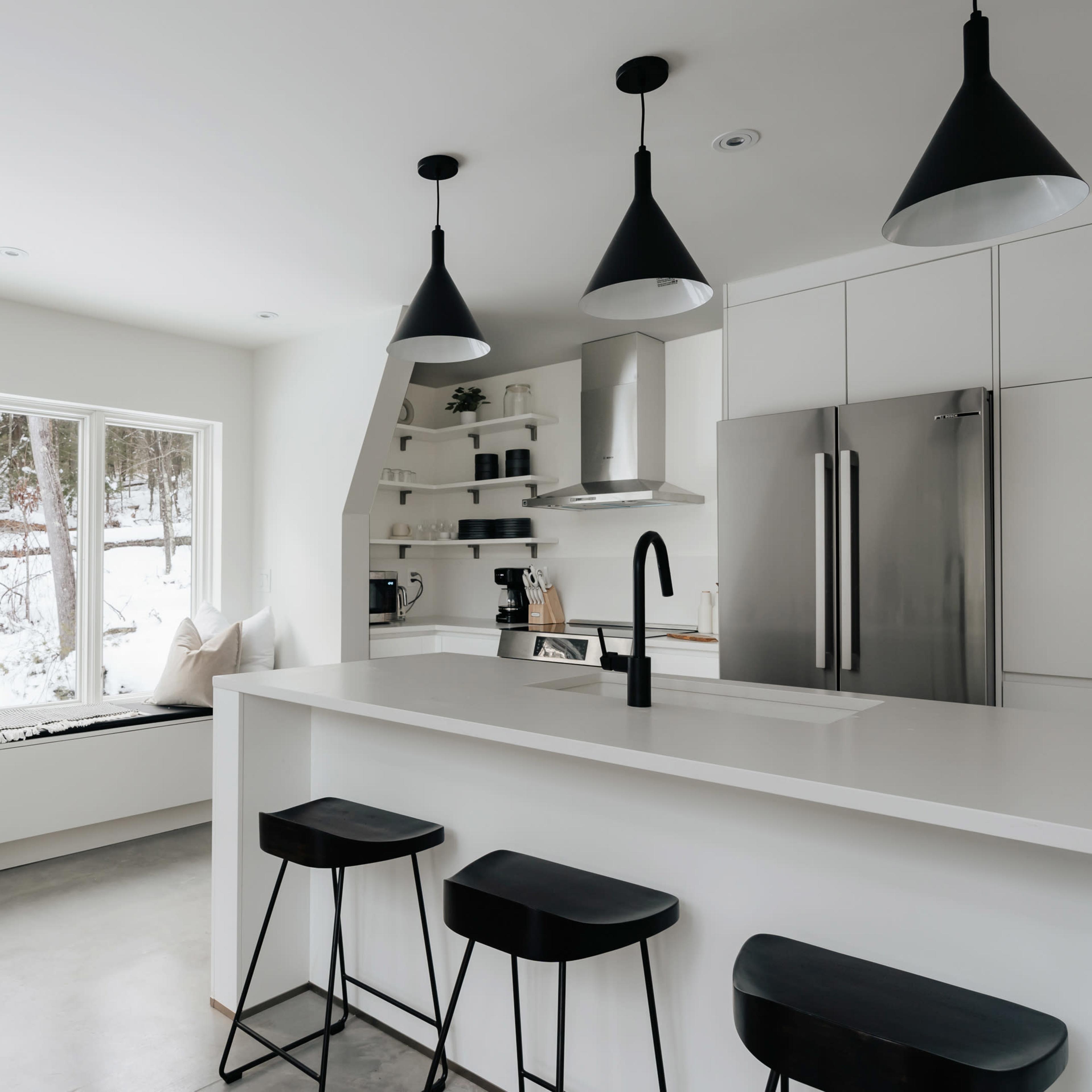 A modern kitchen with a white color scheme, featuring a large window, minimalist shelves, and three black bar stools at a white countertop.