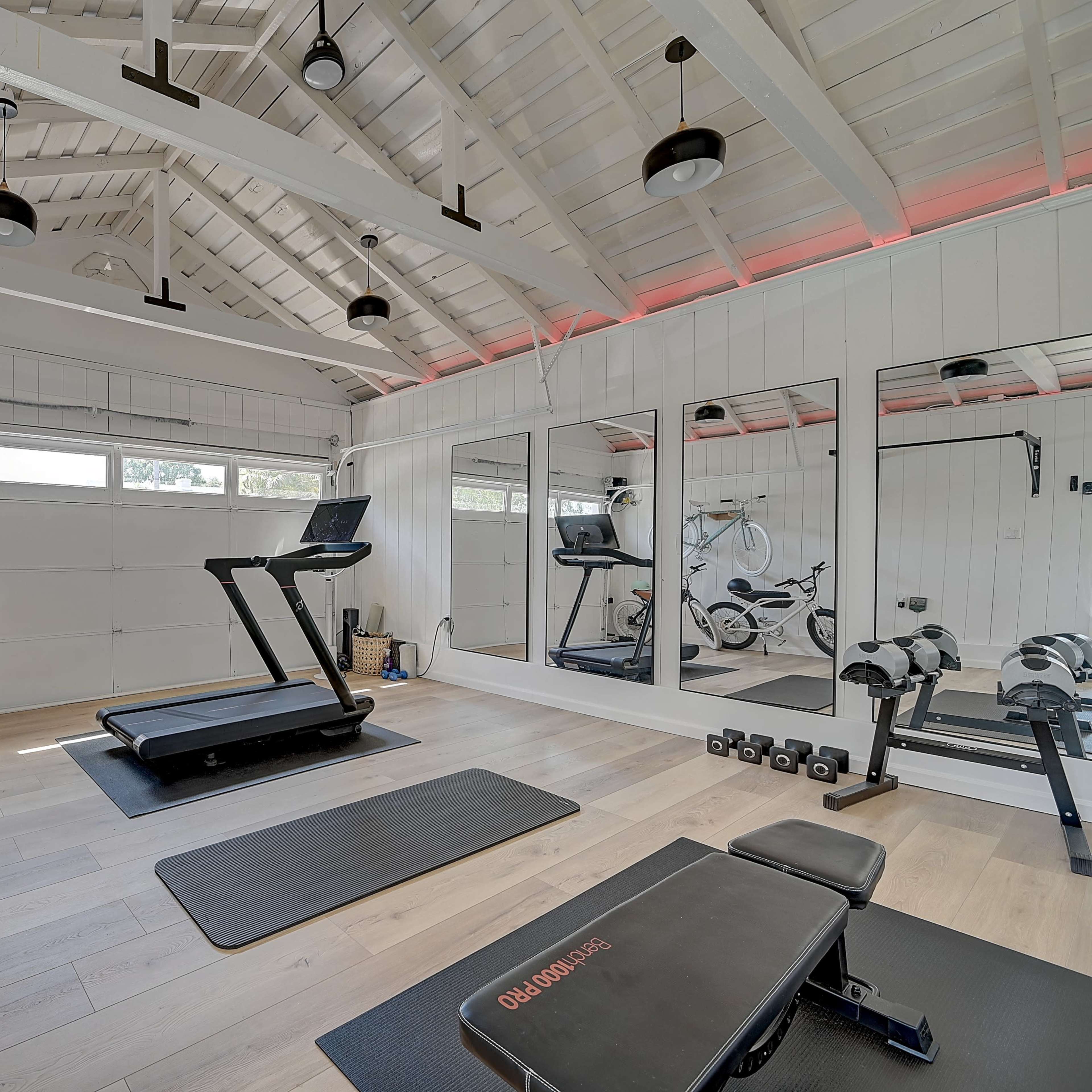 A modern home gym features a treadmill, free weights, and bicycles against a bright, airy backdrop with large mirrors and wooden beams.